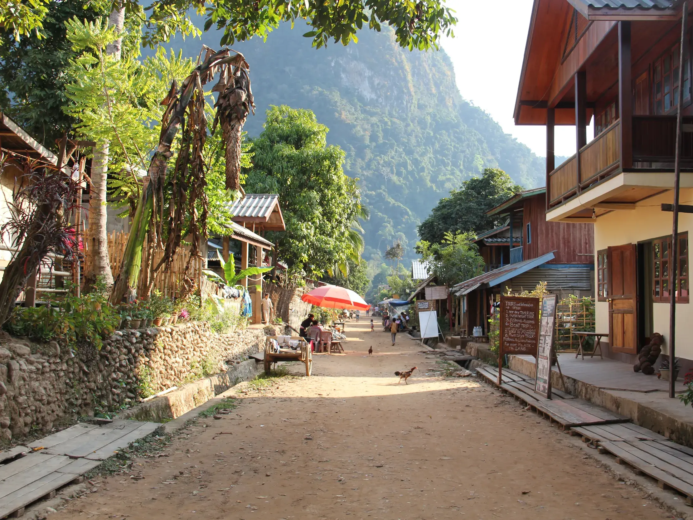 Unbefestigte Dorfstraße in Muang Ngoy, gesäumt von einfachen Holzhäusern vor der Kulisse grüner Karstfelsen in Laos.