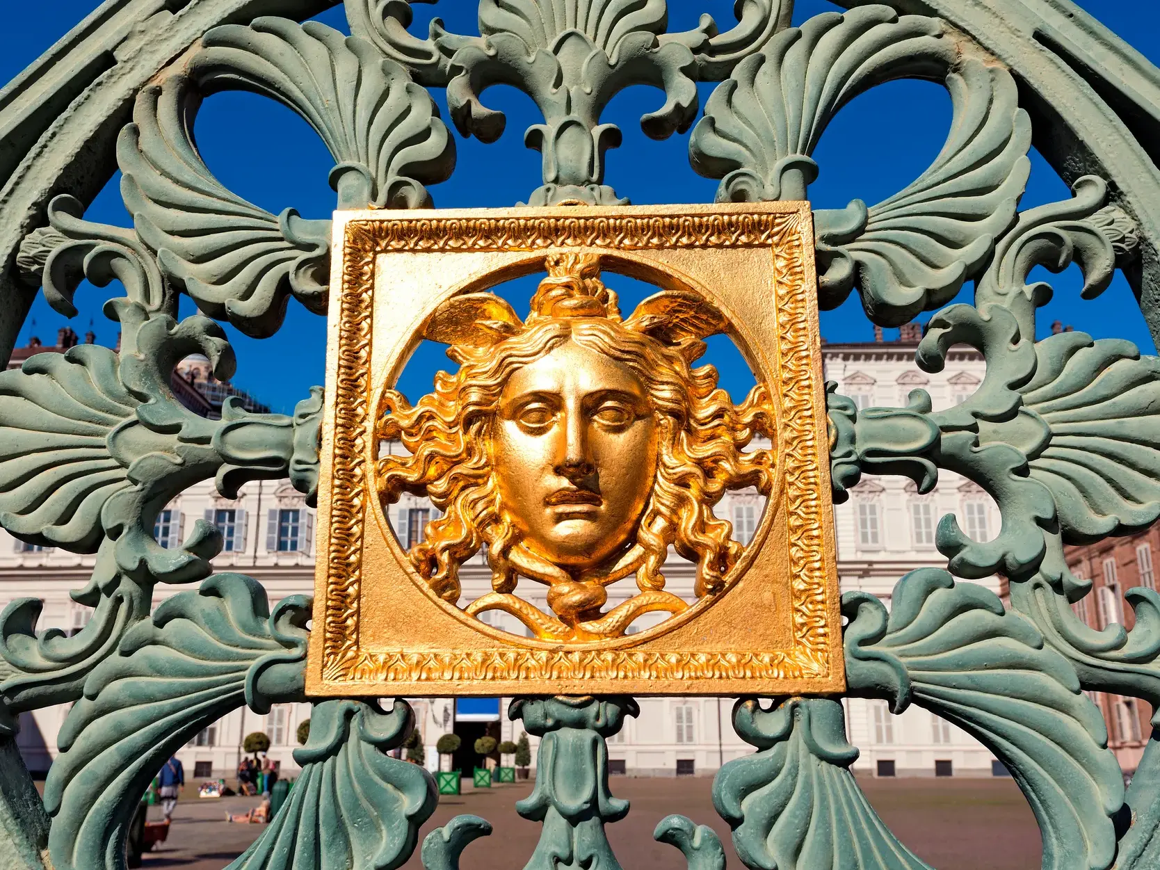 Goldenes Medusenhaupt als Detail am kunstvollen Gitter des Königspalastes in Turin vor einer barocken Fassade.