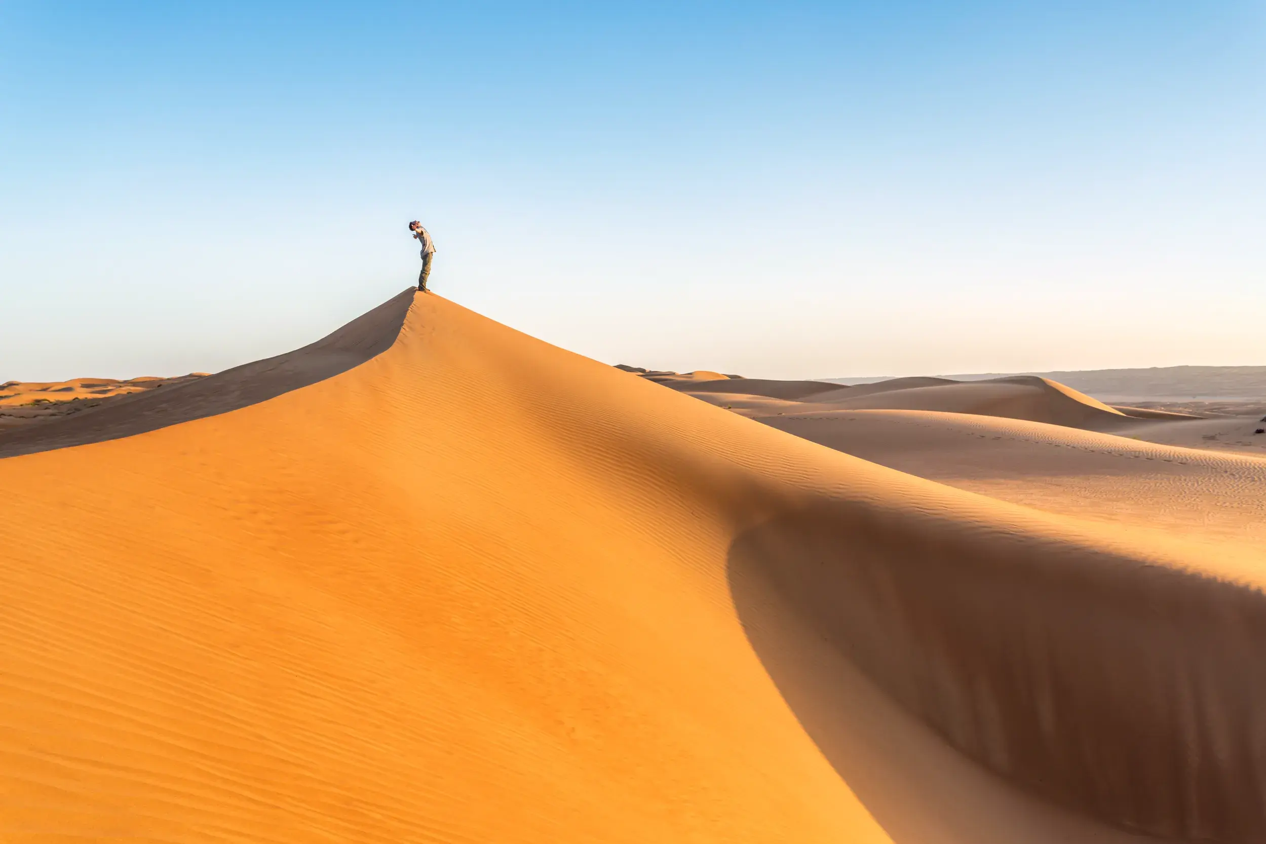Ein Alleinreisender steht auf dem Gipfel einer hohen Sanddüne und blickt bei Sonnenuntergang über die Weite der Wahiba-Wüste im Oman.