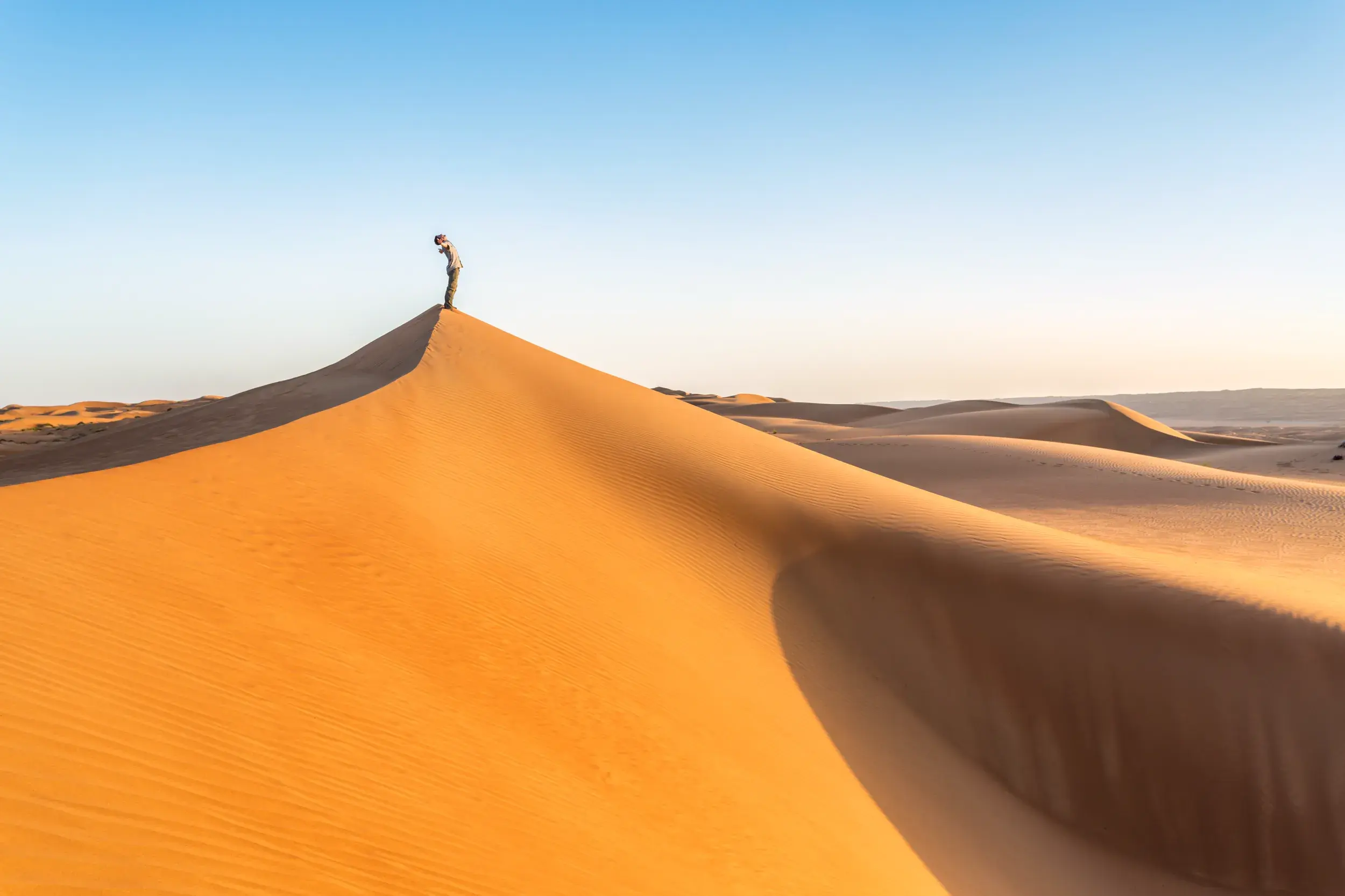Ein Alleinreisender steht auf dem Gipfel einer hohen Sanddüne und blickt bei Sonnenuntergang über die Weite der Wahiba-Wüste im Oman.