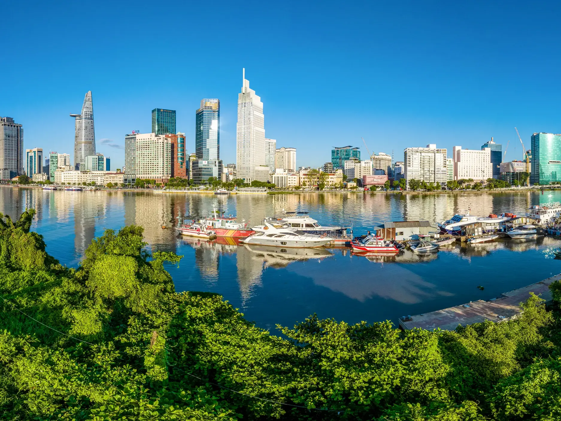 Sonniger Blick über grüne Bäume auf die beeindruckende Skyline von Saigon mit Wolkenkratzern und Booten am Flussufer.