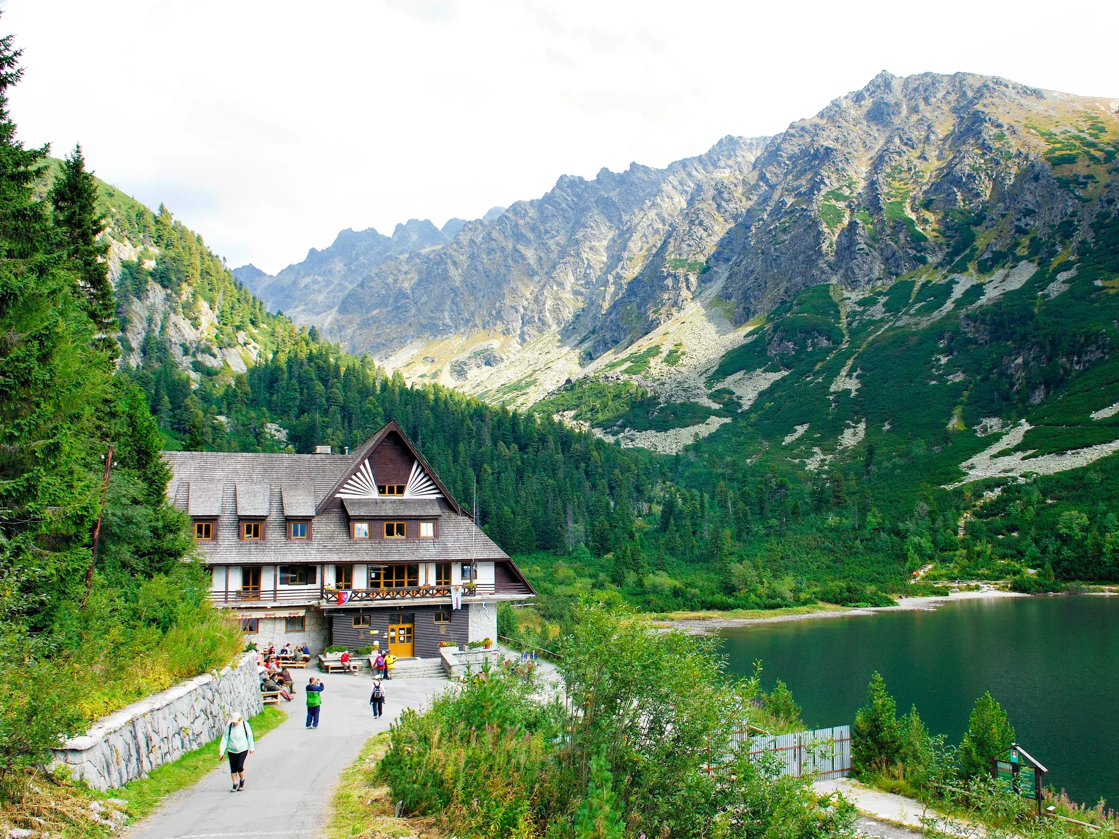Wanderer auf einem Weg zur Berghütte am Ufer des Popradske Pleso Sees vor der eindrucksvollen Bergkulisse der Hohen Tatra.