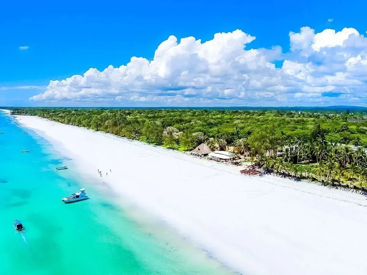 Luftaufnahme vom weißen Sandstrand des Diani Beach in Kenia mit türkisem Wasser, Booten und sattgrünen Palmenwäldern.