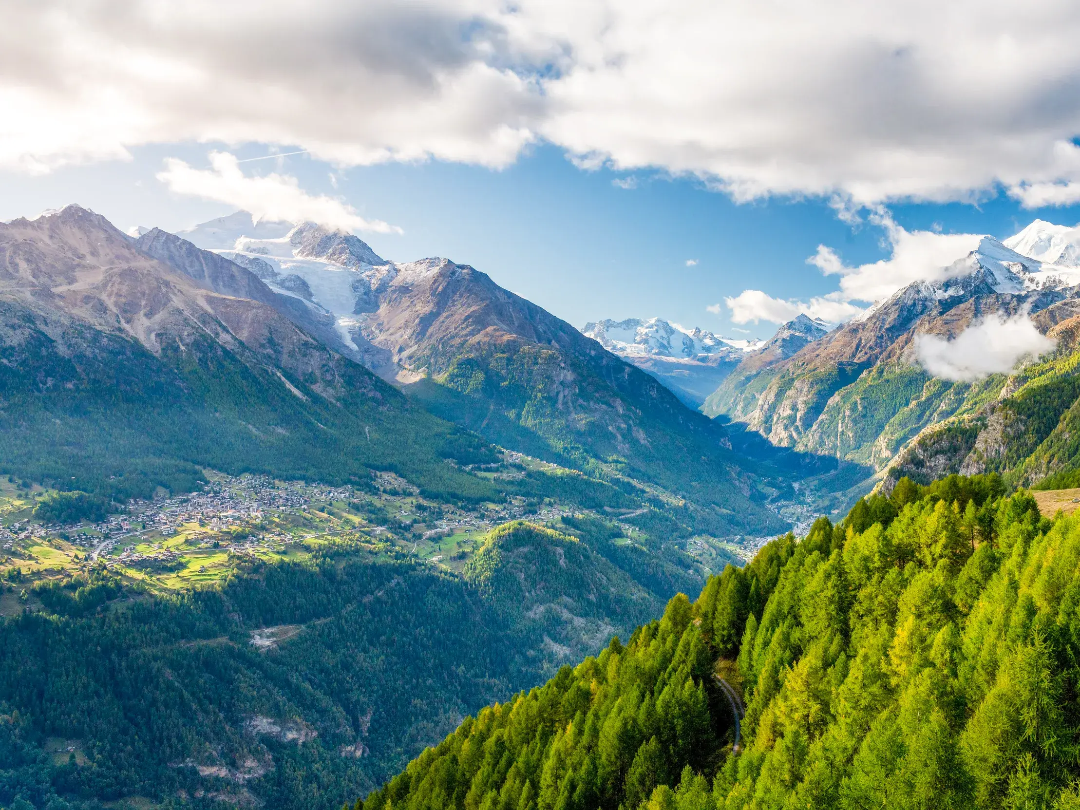 Panoramablick beim Wandern im Mattertal auf grüne Wälder, ein Dorf und die gletscherbedeckten Gipfel der Walliser Bergwelt.