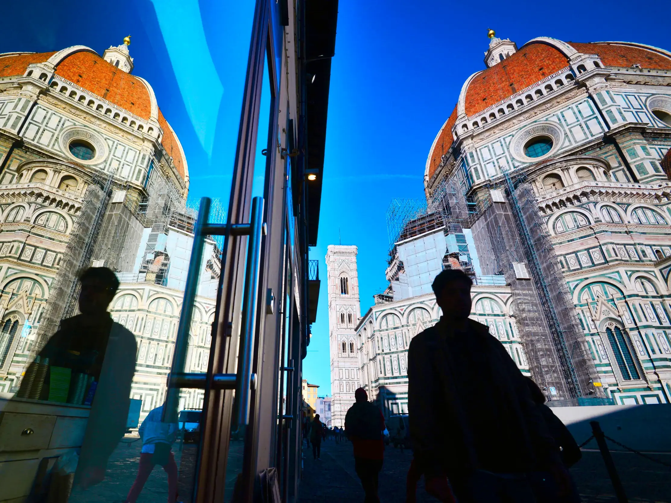 Die berühmte Kuppel des Doms von Florenz spiegelt sich in einer Glasfassade, davor die Silhouetten von Passanten.
