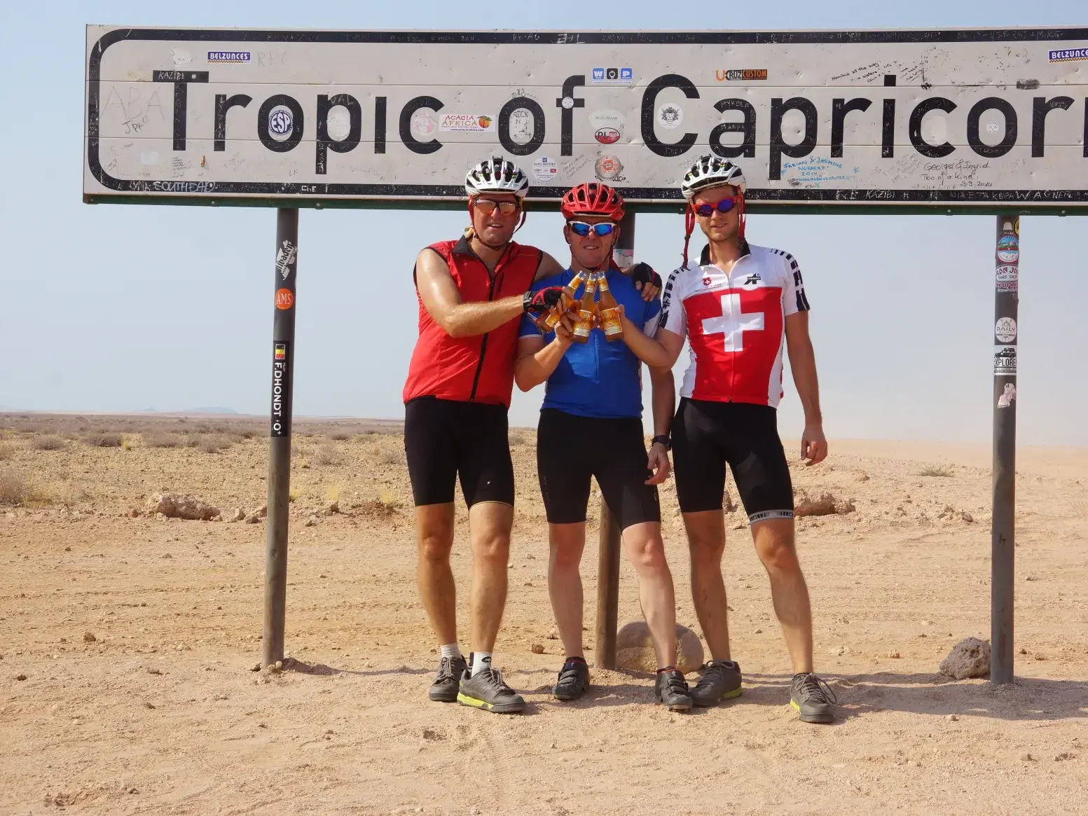 Drei Radfahrer in der Namib-Wüste posieren für ein Foto vor dem Schild des Wendekreises des Steinbocks und stoßen an.