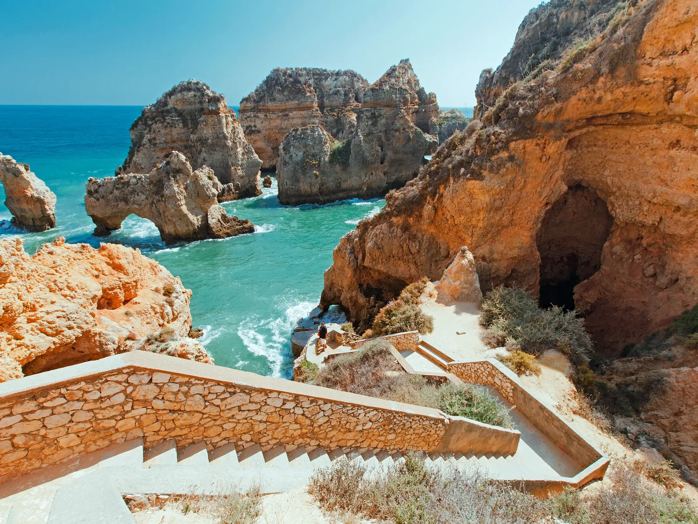 Eine Steintreppe führt an der sonnigen Felsenküste bei Lagos hinab zum türkisen Atlantik mit seinen berühmten Felsformationen.