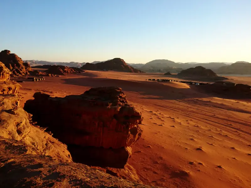 Weite, rote Wüstenlandschaft im Wadi Rum mit einem Zeltlager eingebettet zwischen Sandsteinfelsen im warmen Abendlicht.