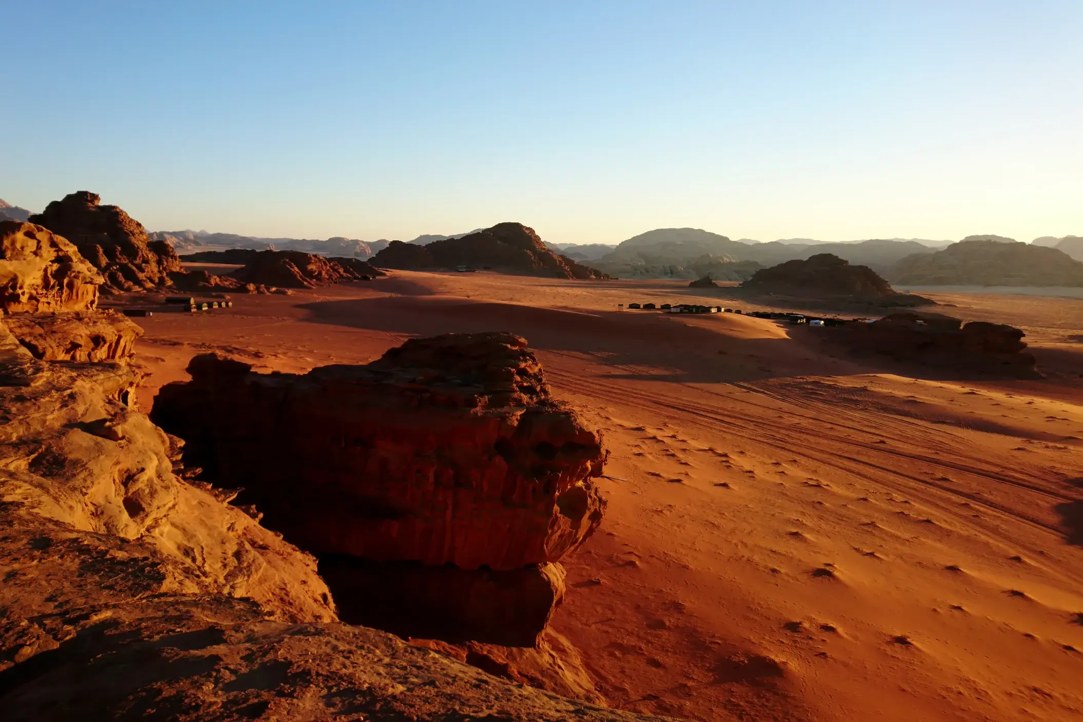 Weite, rote Wüstenlandschaft von Wadi Rum bei Sonnenuntergang mit Blick auf ein Trekking-Camp für Wanderer in der Ferne.