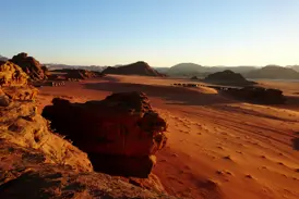 Weite, rote Wüstenlandschaft von Wadi Rum bei Sonnenuntergang mit Blick auf ein Trekking-Camp für Wanderer in der Ferne.
