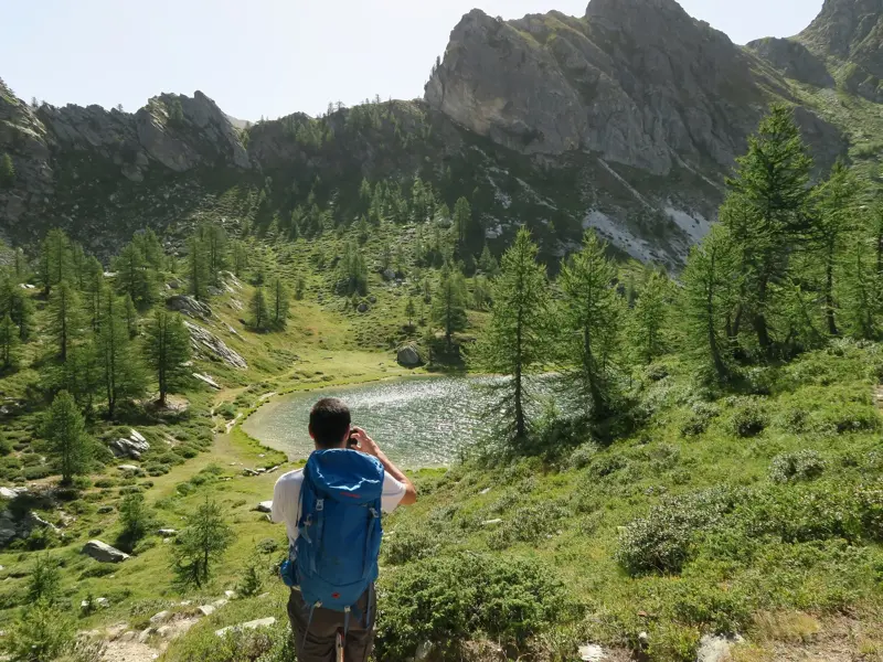 Wanderer in Rückansicht fotografiert einen glitzernden Bergsee umgeben von Lärchen und Felsgipfeln im Maira-Tal.