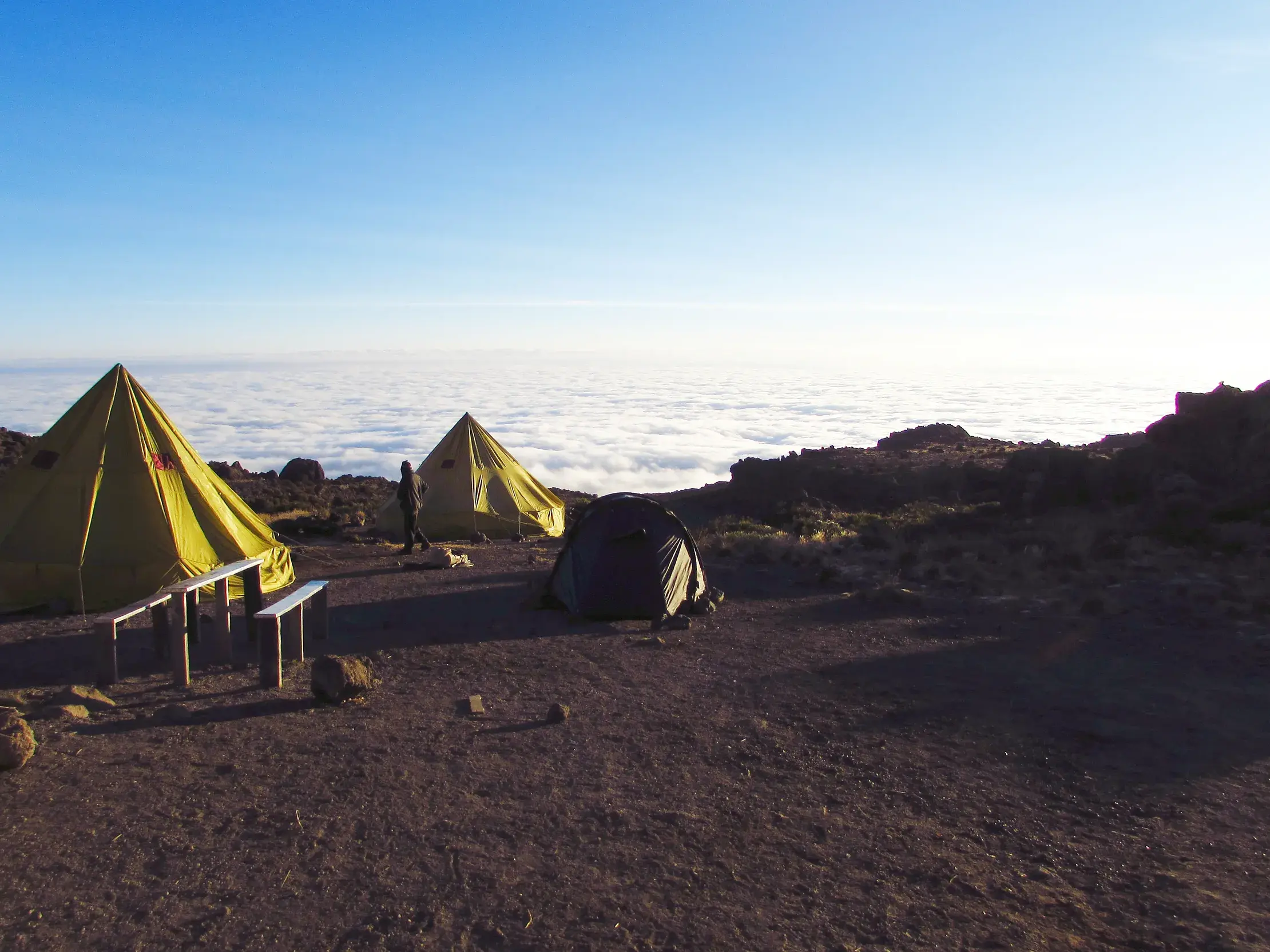 Ein Trekking-Camp mit gelben Zelten in der kargen Landschaft des Kilimanjaro, hoch über einem geschlossenen Wolkenmeer.