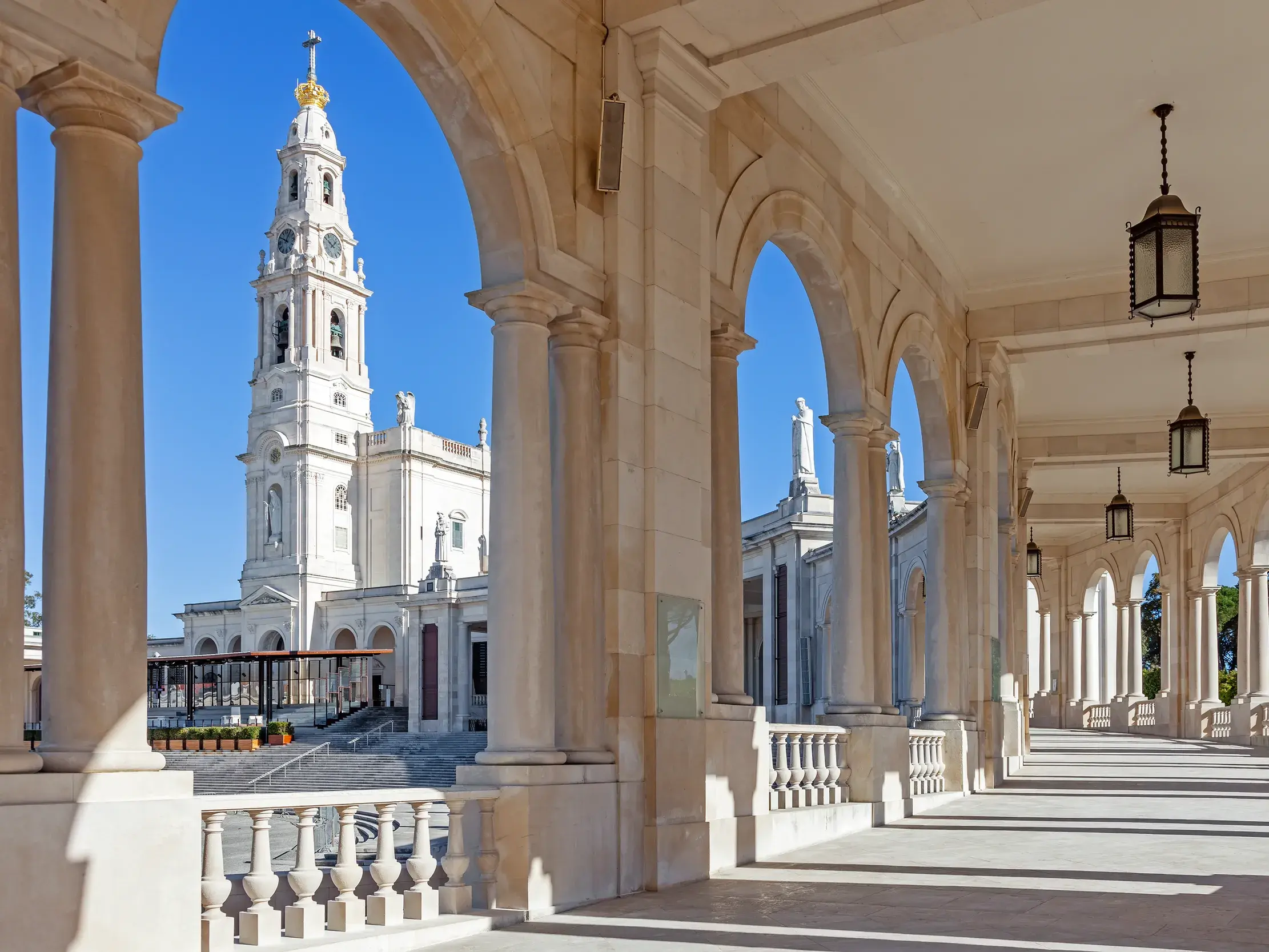 Blick durch den sonnigen Säulengang der Kolonnaden auf die imposante Rosenkranzbasilika im Wallfahrtsort Fátima.
