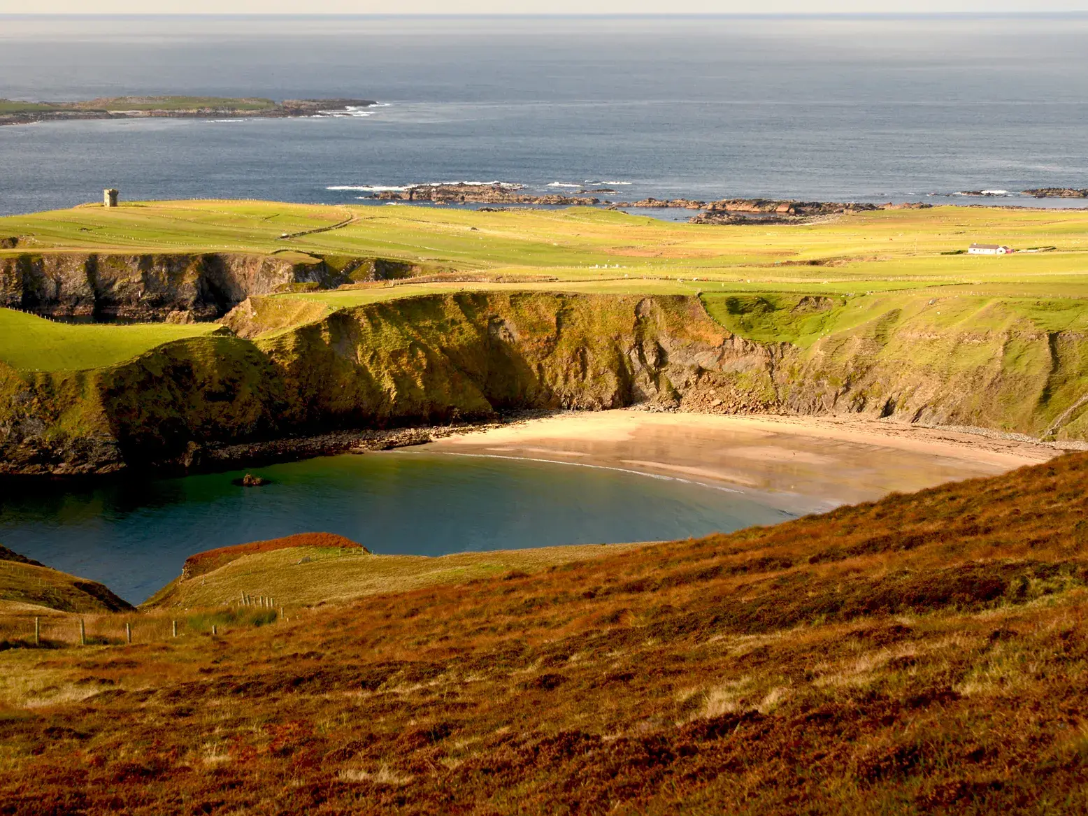 Blick von einem heidebewachsenen Hügel auf eine Bucht mit Sandstrand und die Steilklippen bei Glencolumbkille in Irland.