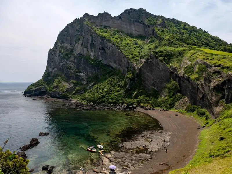 Blick auf den grün bewachsenen Tuffkegel Seongsan Ilchulbong auf Jeju mit einer Bucht mit schwarzem Strand und türkisfarbenem Wasser.