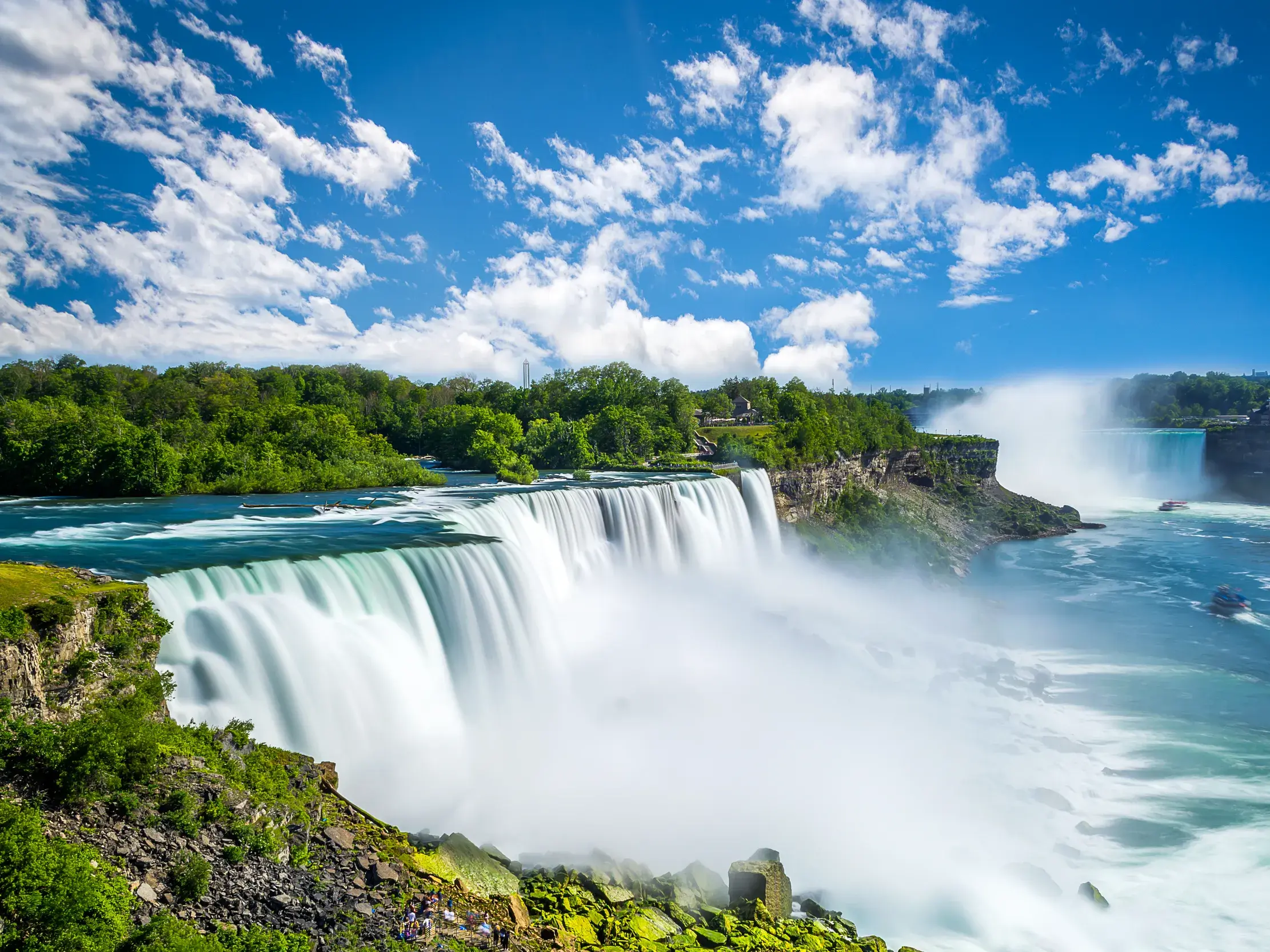 Die gewaltigen Niagarafälle in Kanada an einem sonnigen Tag. Ausflugsboote fahren in die aufsteigende Gischt.