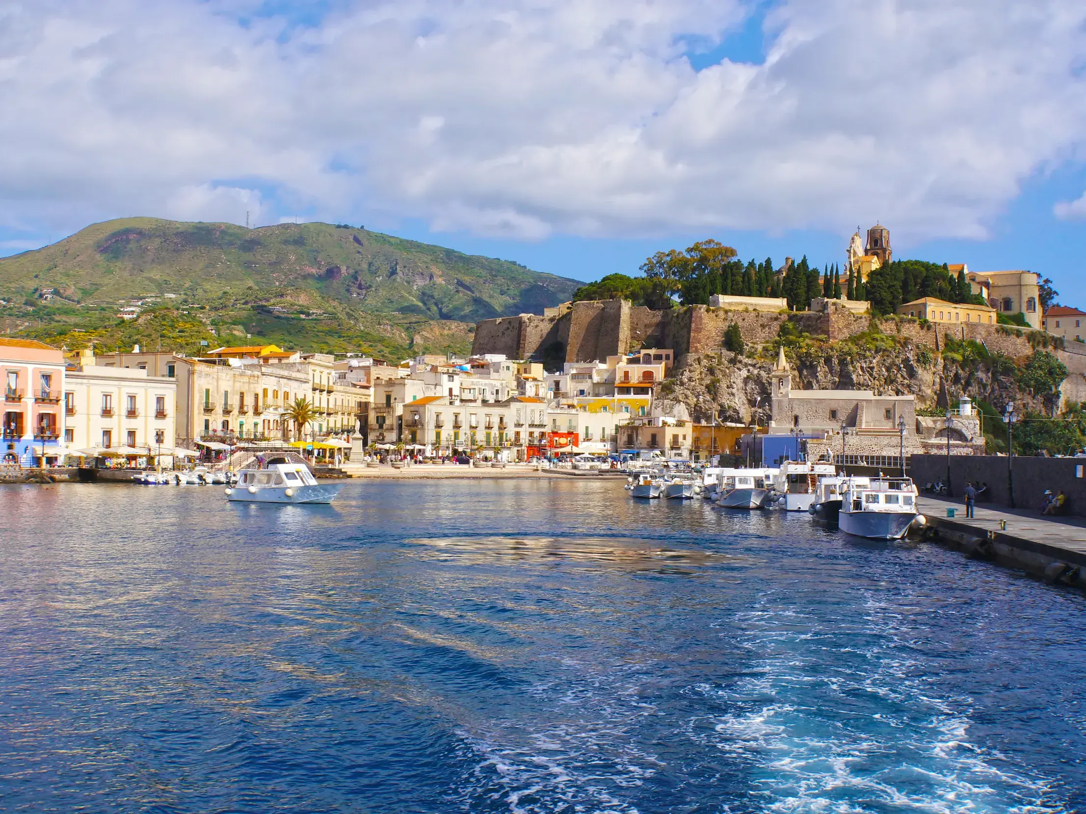 Malerischer Blick vom Meer auf den Hafen von Lipari mit dem Castello und bunten Häusern vor einem grünen Berg.