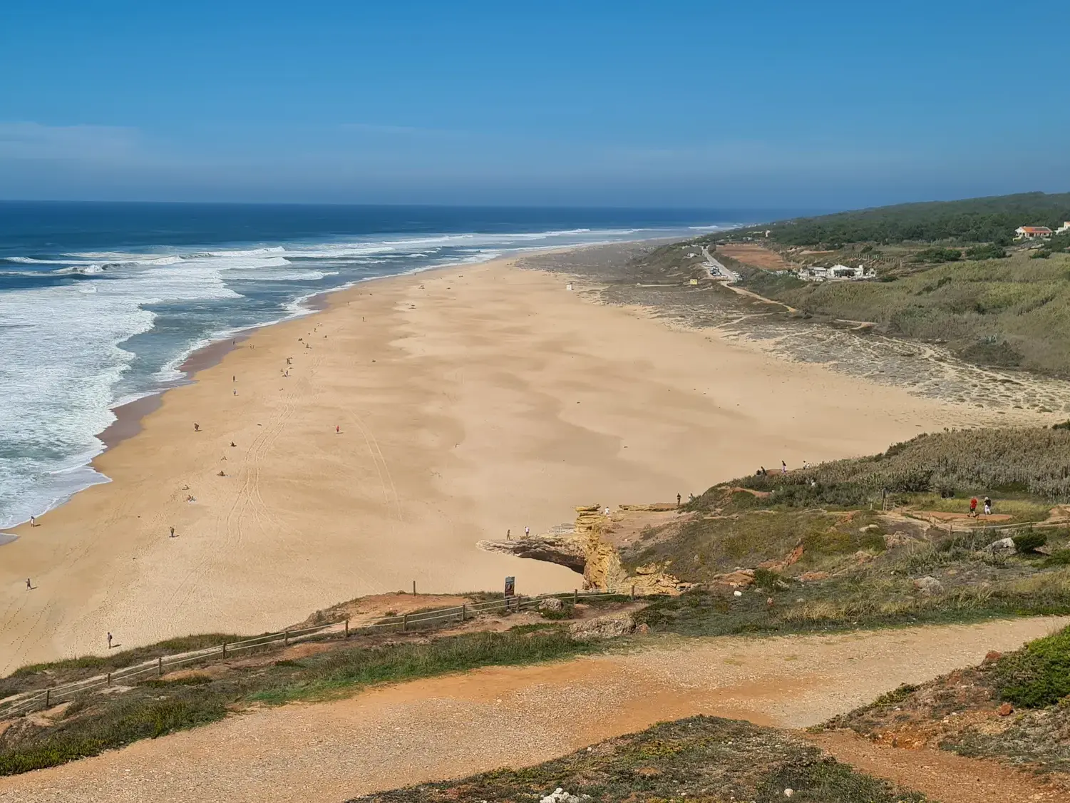 Wanderweg entlang der Klippen mit Panoramablick auf den weiten Sandstrand und die Wellen des Atlantiks bei Nazaré.