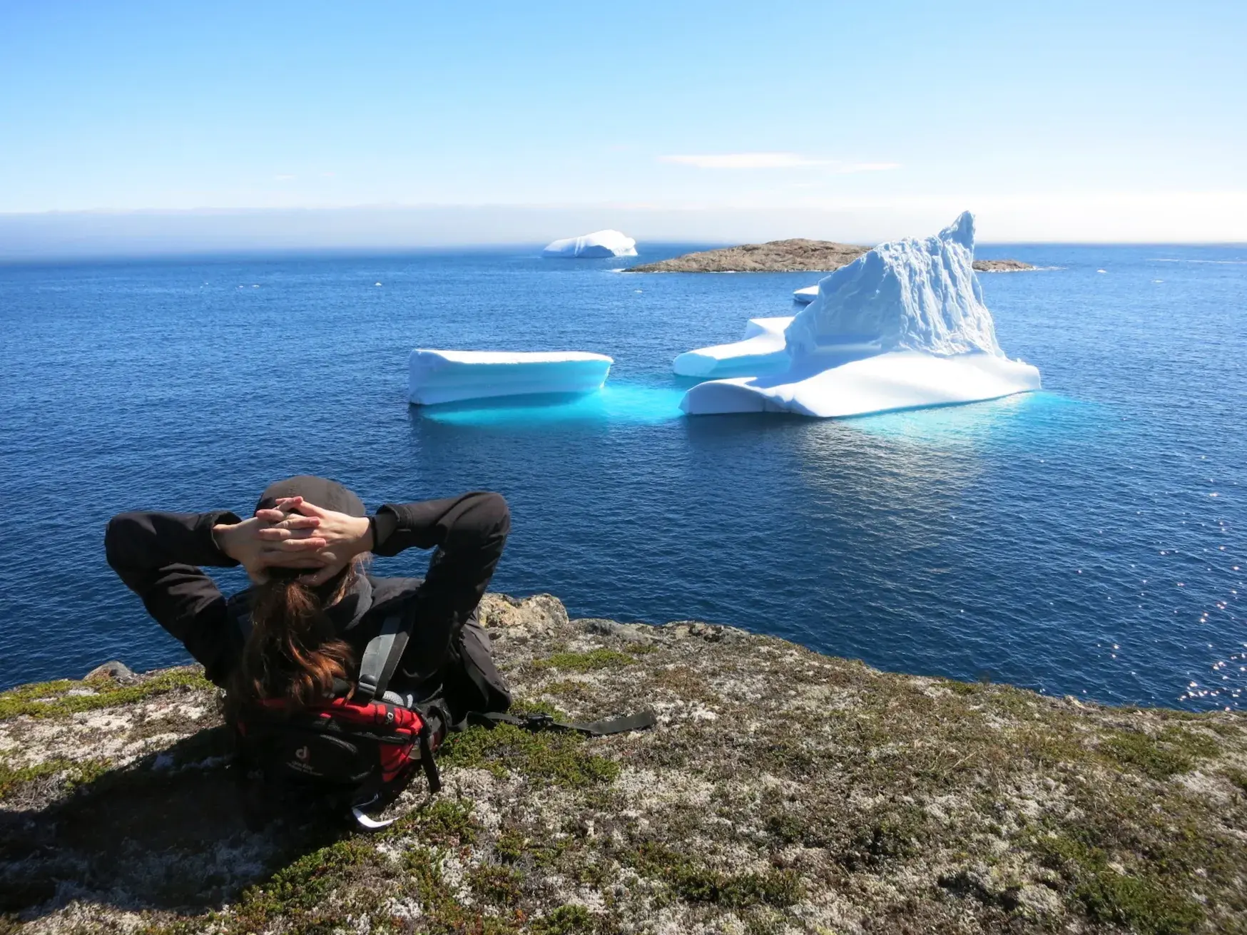 Eine Wanderin entspannt auf einem moosbewachsenen Felsen und blickt auf riesige Eisberge im blauen Polarmeer Ostgrönlands.