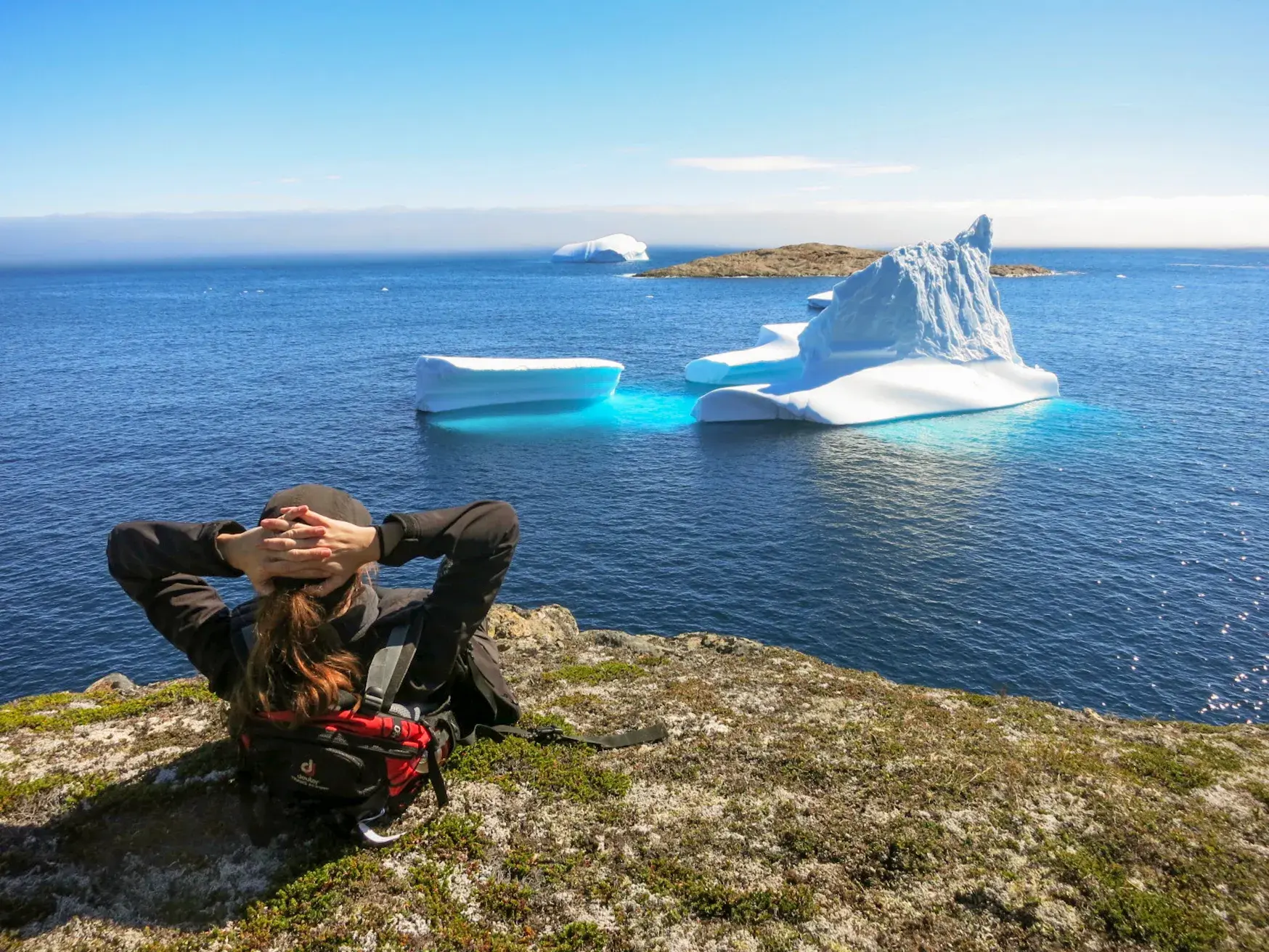 Eine Wanderin macht Pause auf einer Klippe in Ostgrönland und blickt auf gigantische Eisberge im blauen Polarmeer.