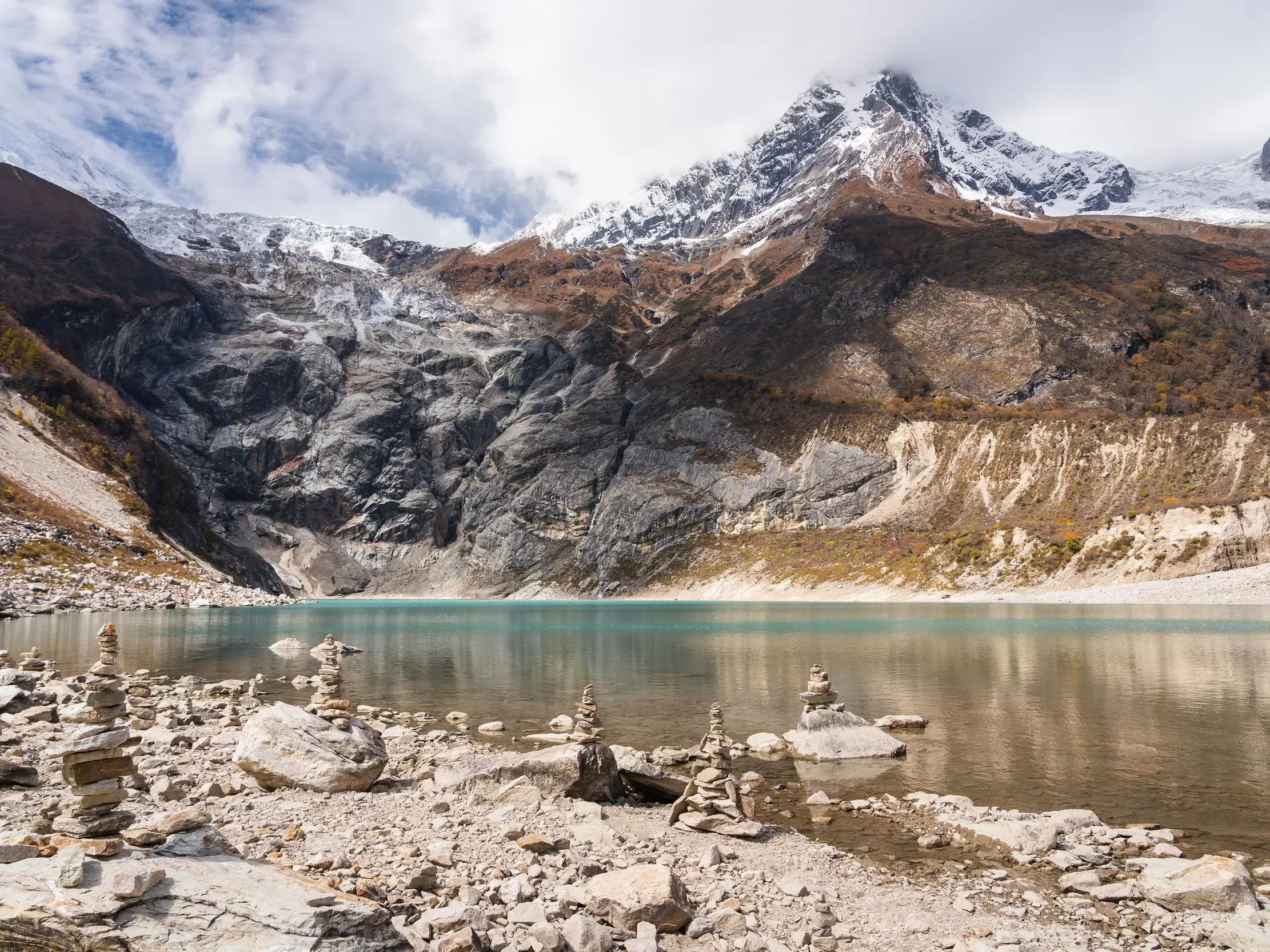 Steinerne Ufer des türkisen Birendra-Sees in Nepal mit Steinmännchen und Blick auf das schneebedeckte Manaslu-Massiv.