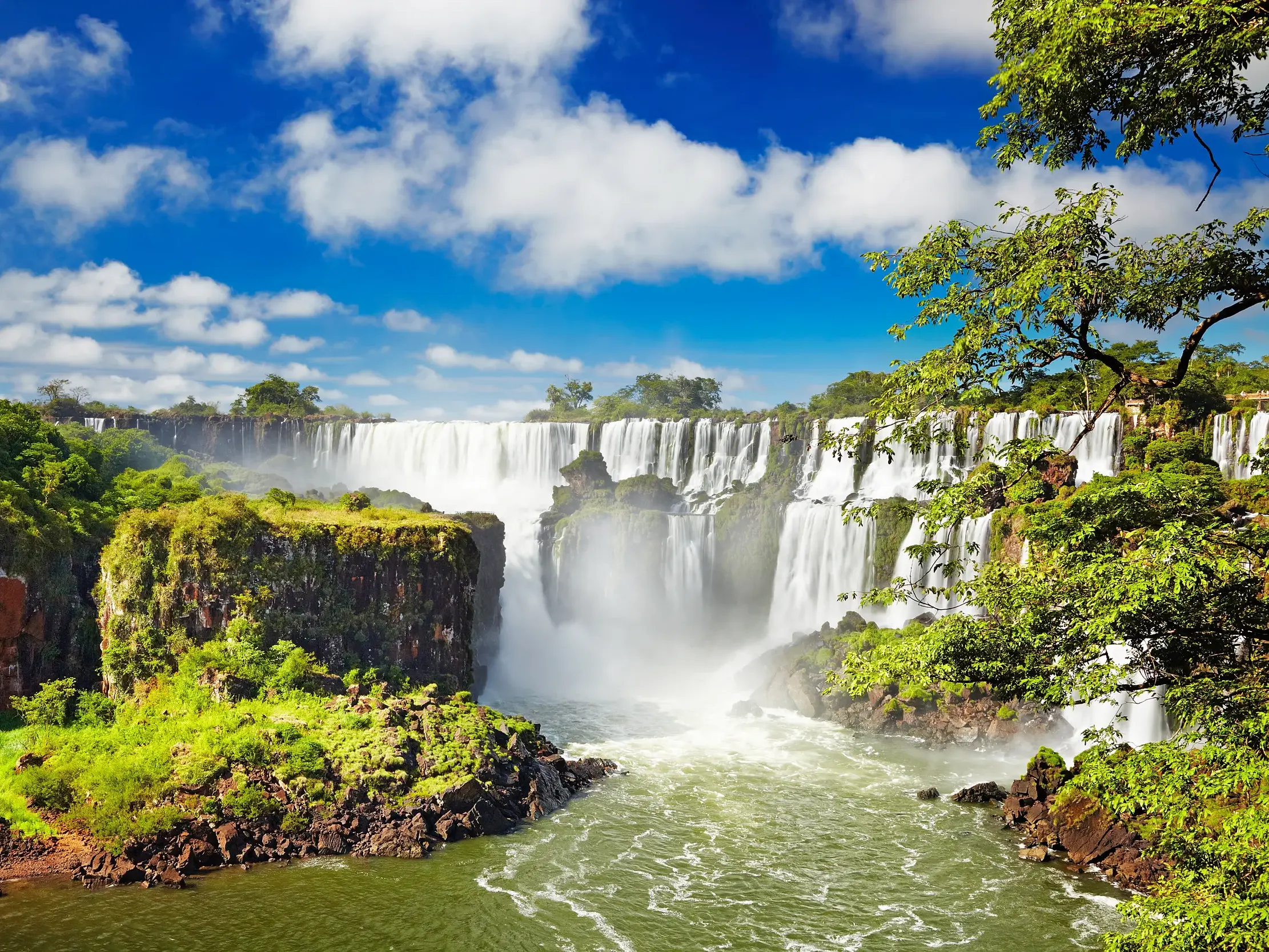 Die mächtigen Iguazú-Wasserfälle (UNESCO-Welterbe) stürzen umgeben von üppigem Regenwald bei strahlend blauem Himmel in die Tiefe.