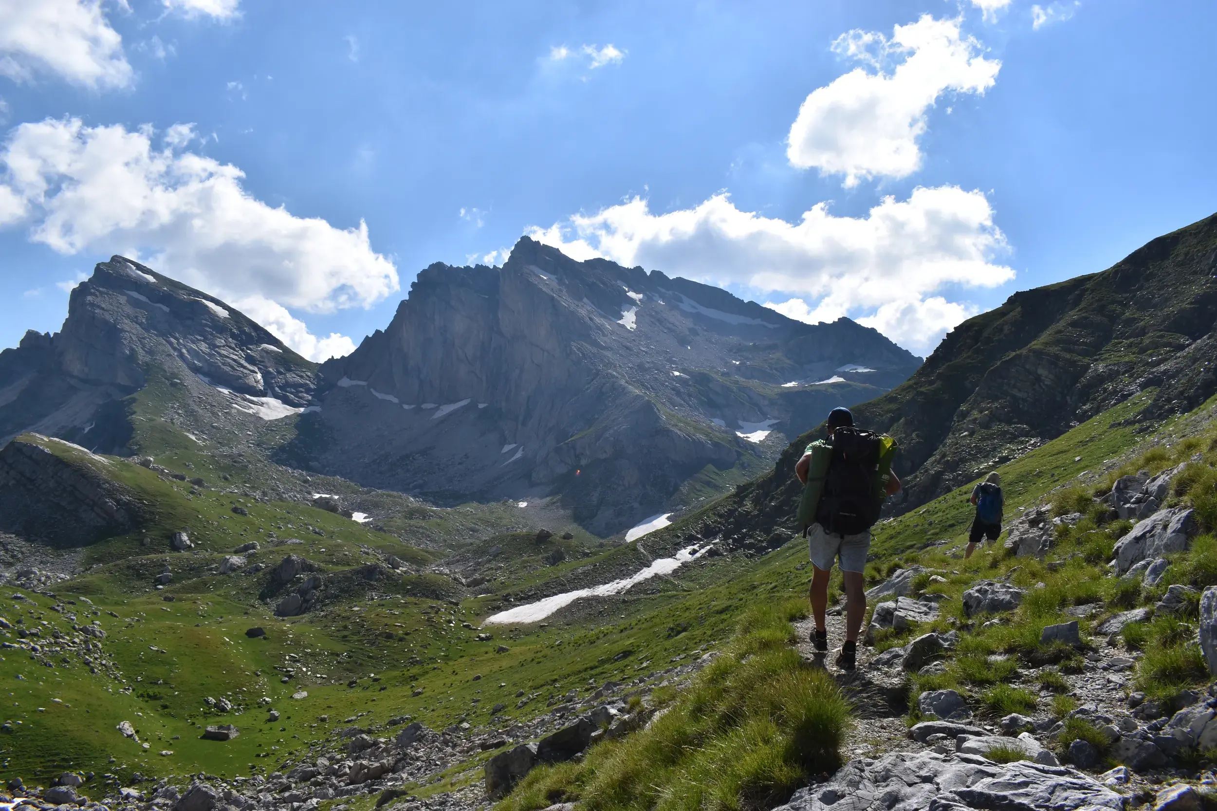 Zwei Trekker mit großen Rucksäcken wandern auf einem schmalen Pfad durch die grüne Berglandschaft des Korab-Gebirges in Albanien.