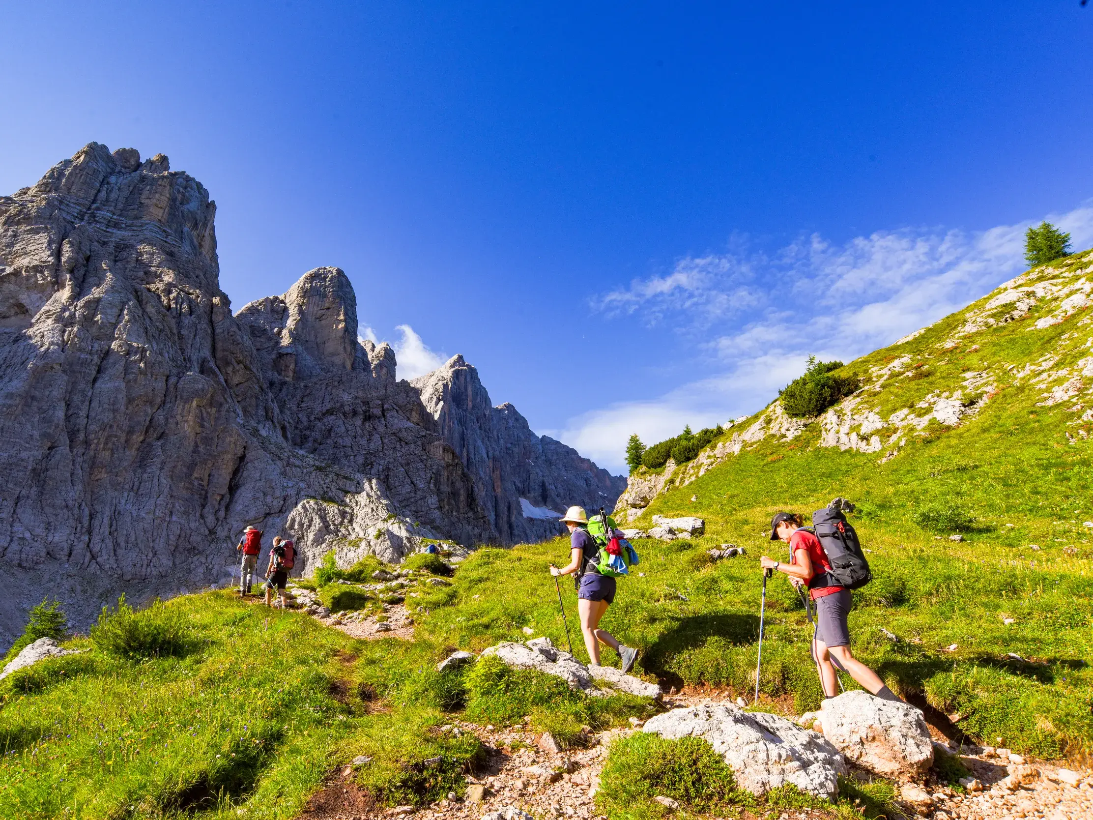 Wandergruppe mit Rucksäcken auf einem schmalen Pfad des Höhenwegs Alta Via 1 entlang schroffer Felsen der Dolomiten.