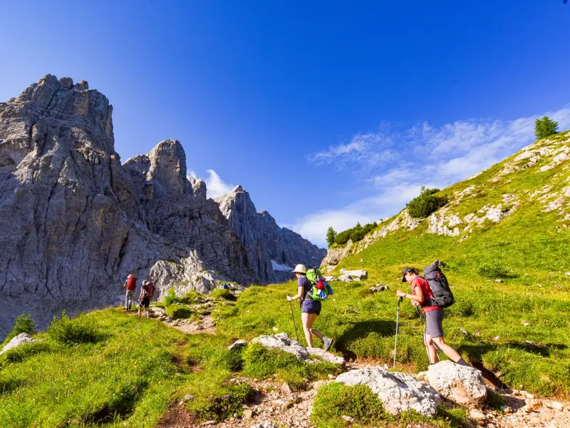 Wandergruppe mit Rucksäcken auf einem schmalen Pfad des Höhenwegs Alta Via 1 entlang schroffer Felsen der Dolomiten.
