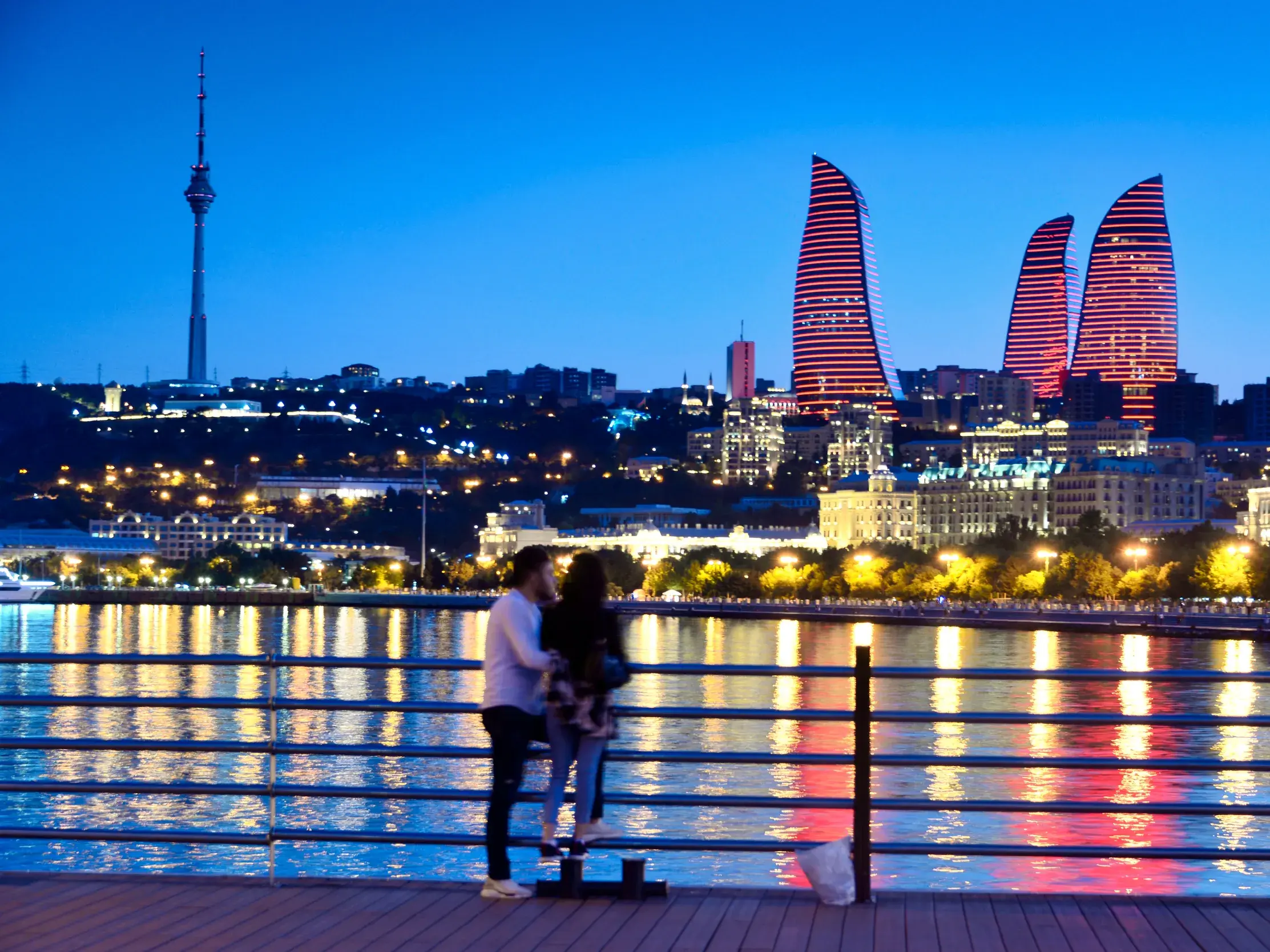 Paar blickt von der Uferpromenade auf die Skyline von Baku mit den rot leuchtenden Flame Towers und dem Fernsehturm bei Nacht.