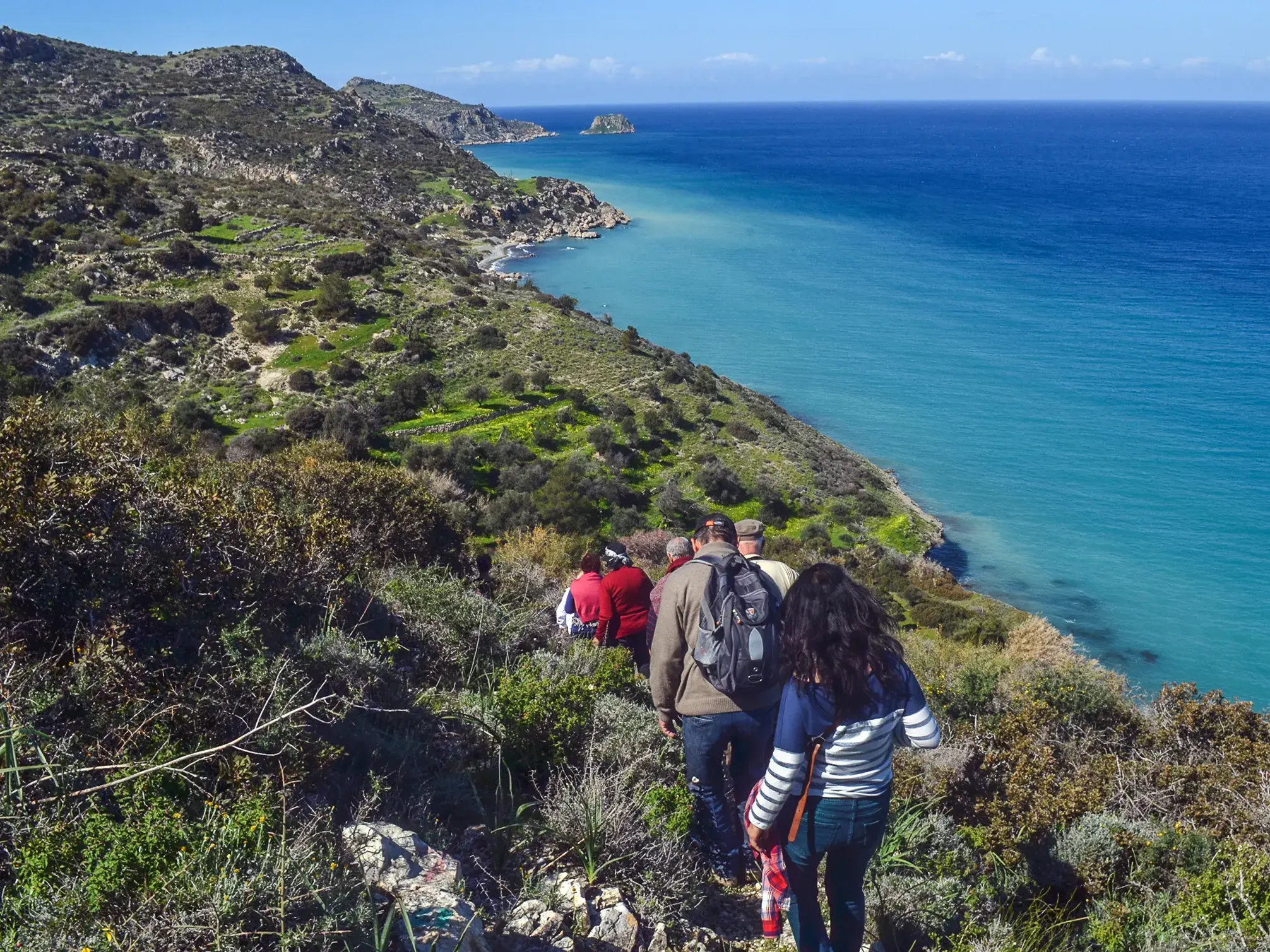 Eine kleine Wandergruppe auf einem Ziegenpfad entlang der grünen Steilküste Nordzyperns mit Blick aufs türkisblaue Meer.