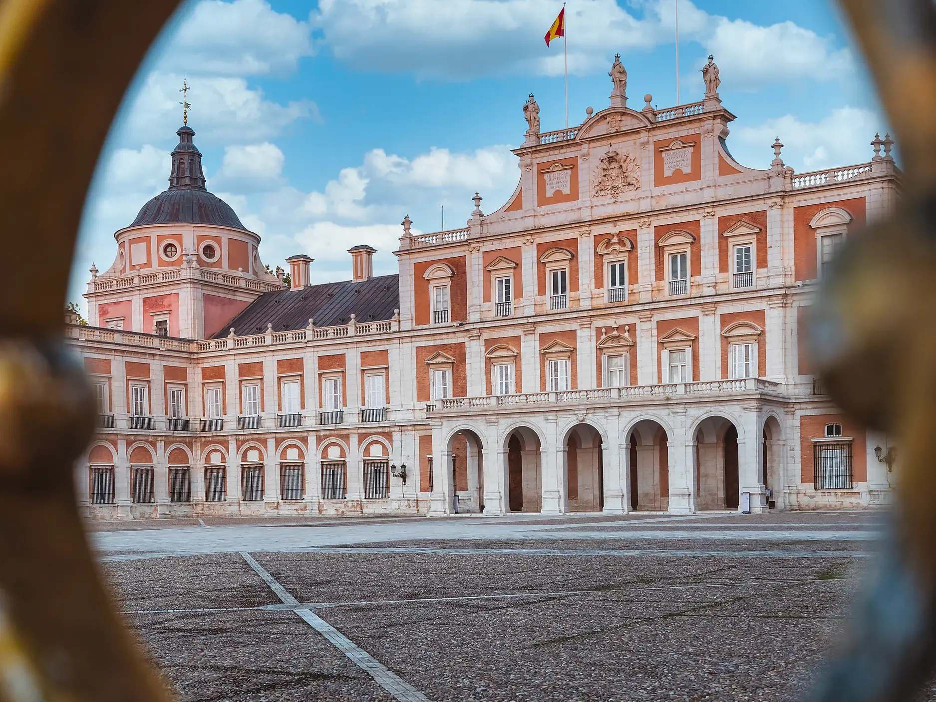 Blick durch ein verziertes Tor auf den großen Vorplatz und die Fassade des heiter-verspielten Sommerschlosses von Aranjuez.