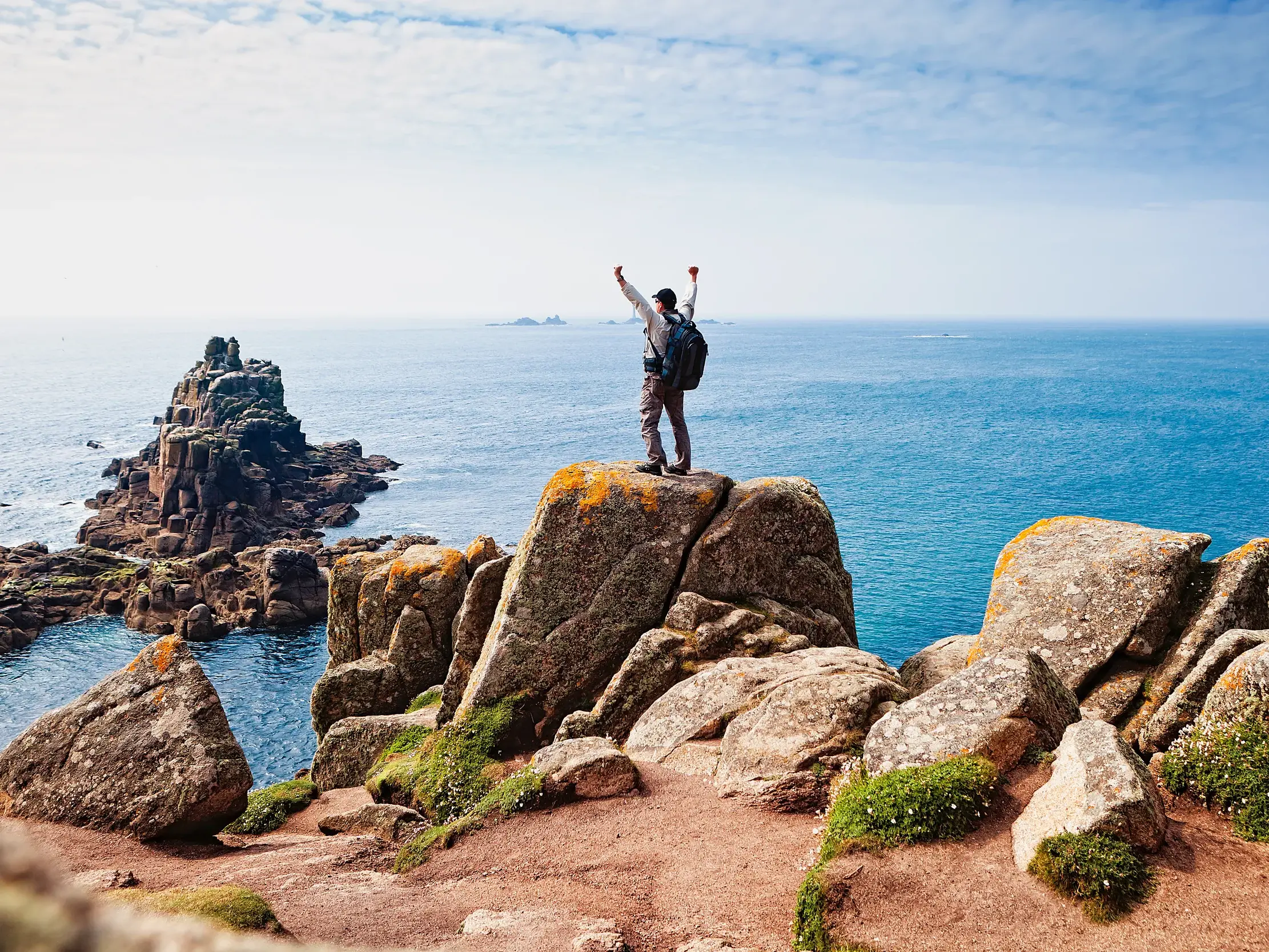 Ein Wanderer mit Rucksack steht mit erhobenen Armen auf einer Felsklippe bei Land's End in Cornwall und blickt auf den Atlantik.