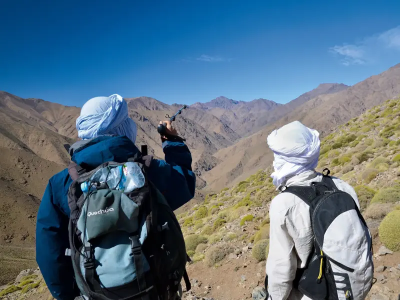 Zwei Wanderer mit Turbanen und Rucksäcken beim Trekking im sonnigen Hohen Atlas mit Blick auf die marokkanische Bergkette.