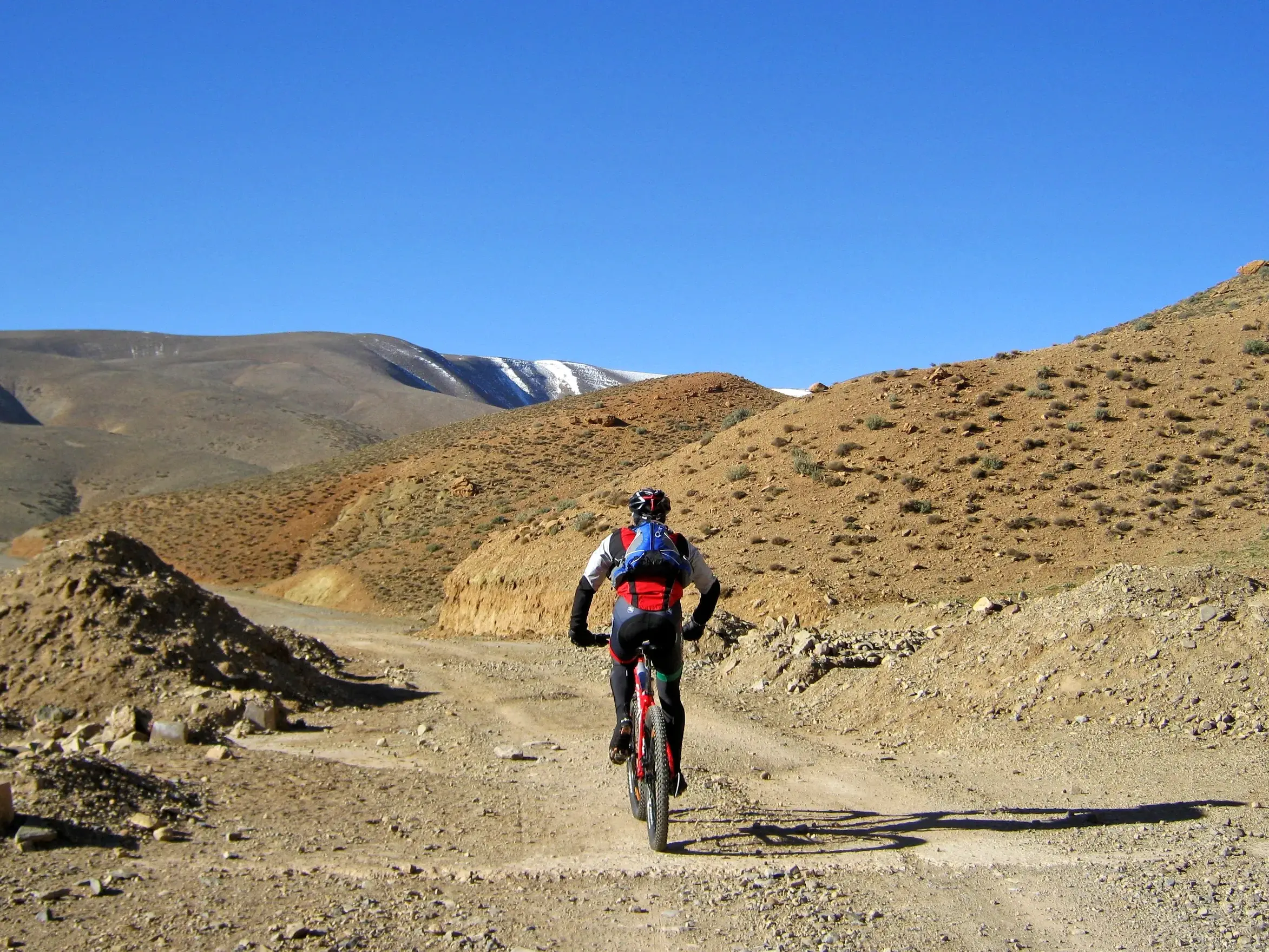 Mountainbiker auf einer Schotterpiste bei der Überquerung des Jebel Sarhro Massivs in Marokko, am Horizont die Gipfel des Hohen Atlas.