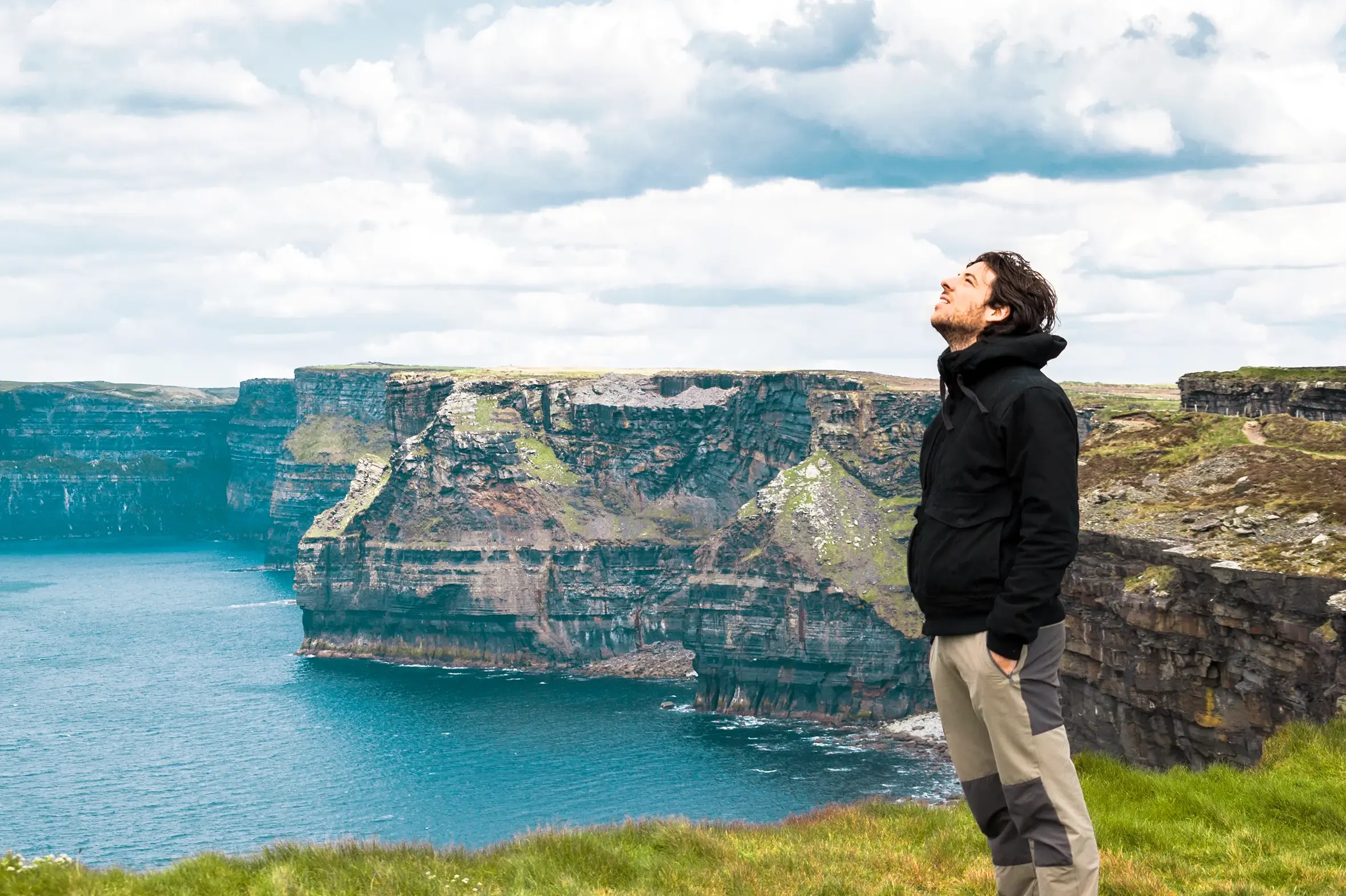 Ein Mann in Wanderkleidung genießt auf einer Irland-Rundreise den weiten Ausblick von den grünen Cliffs of Moher am Atlantik.