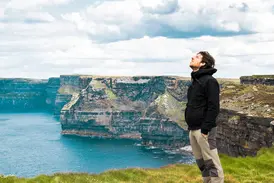 Ein Mann in Wanderkleidung genießt auf einer Irland-Rundreise den weiten Ausblick von den grünen Cliffs of Moher am Atlantik.