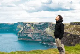 Ein Mann in Wanderkleidung genießt auf einer Irland-Rundreise den weiten Ausblick von den grünen Cliffs of Moher am Atlantik.