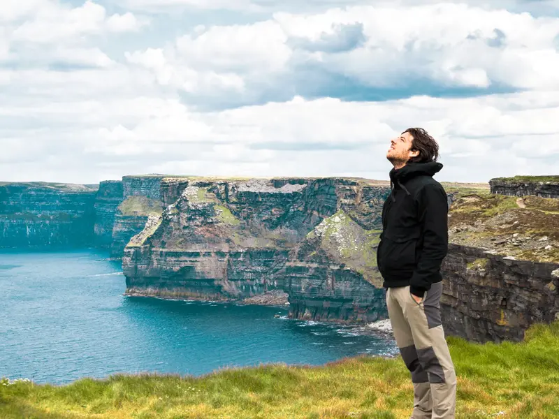 Ein Mann in Wanderkleidung genießt auf einer Irland-Rundreise den weiten Ausblick von den grünen Cliffs of Moher am Atlantik.