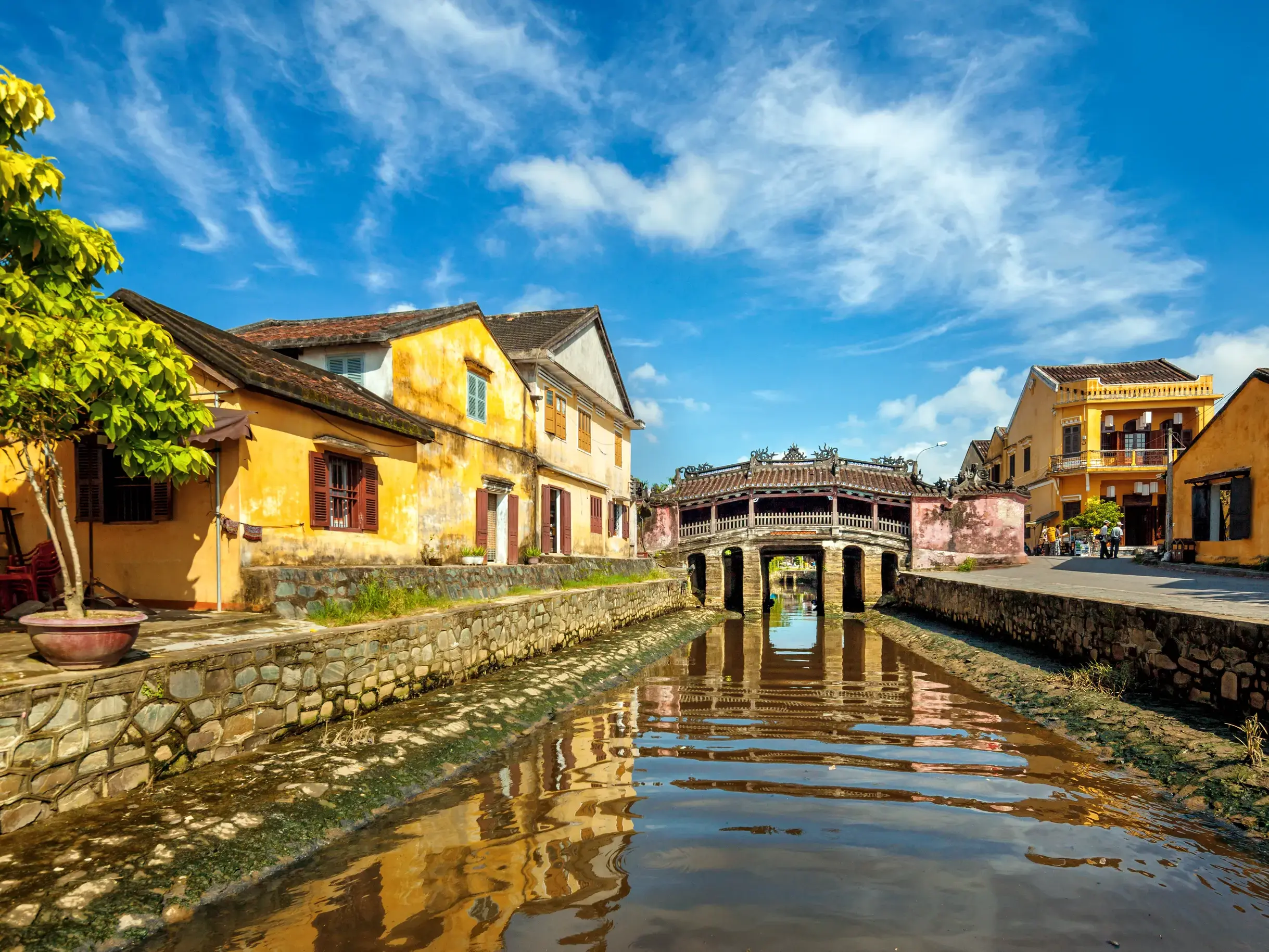 Die berühmte Japanische Brücke in der Altstadt von Hoi An spiegelt sich mit gelben Häusern in einem Kanal wider.