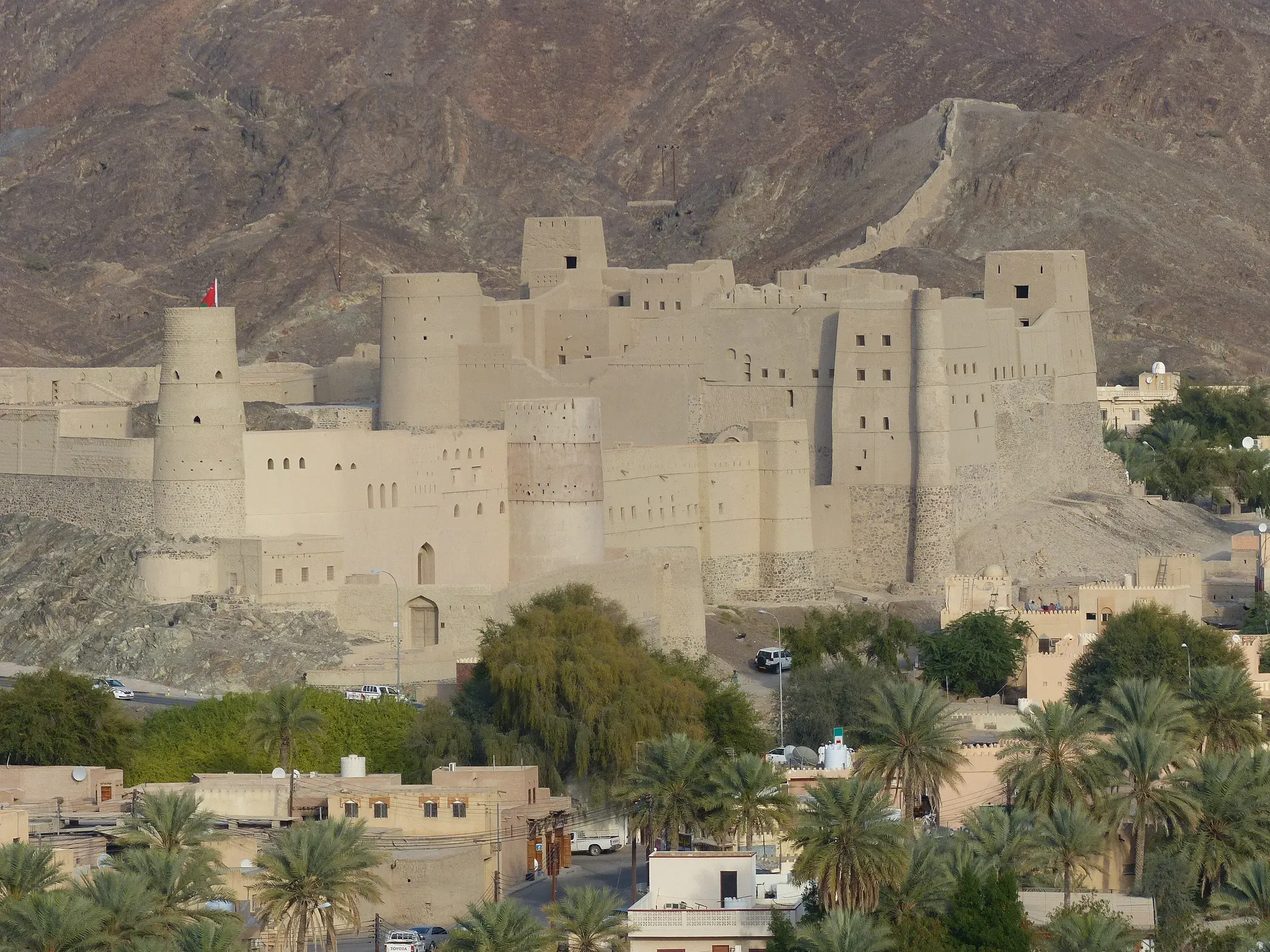 Mächtige Lehmwände der Festung Bahla im Oman mit Palmenhain vor dem schroffen Hadschar-Gebirge.