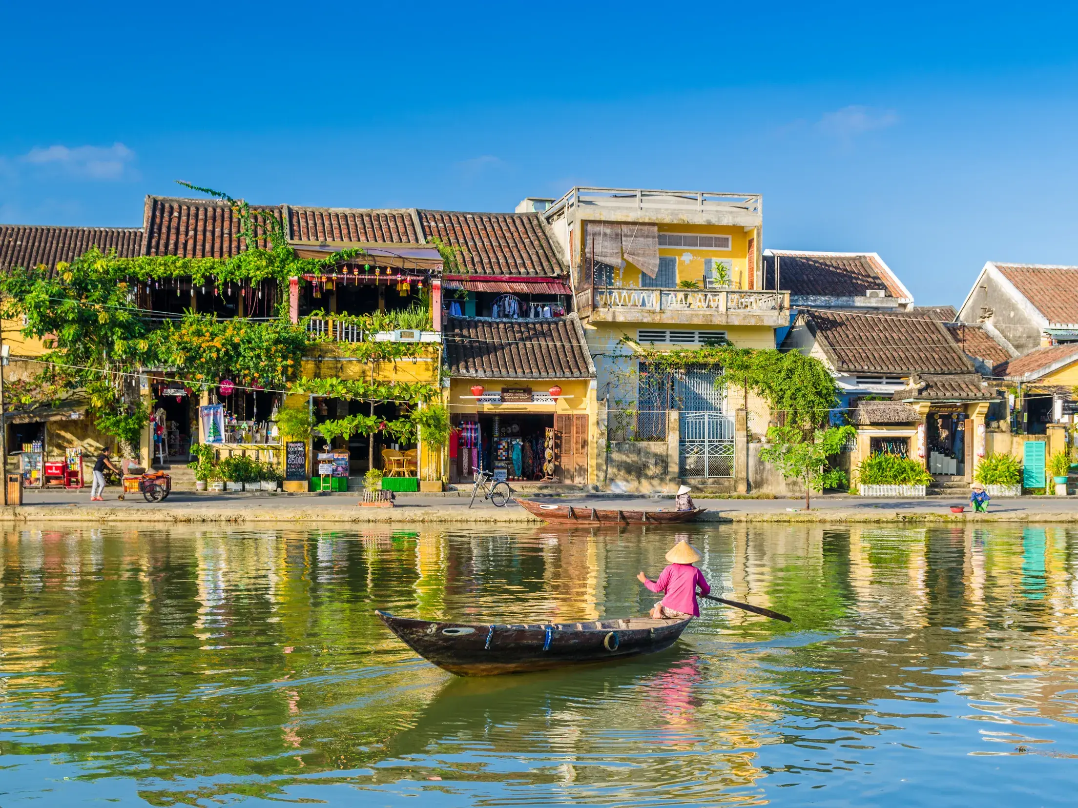 Einheimische Person rudert in einem Holzboot auf dem Fluss entlang der gelben Fassaden der Altstadt von Hoi An.