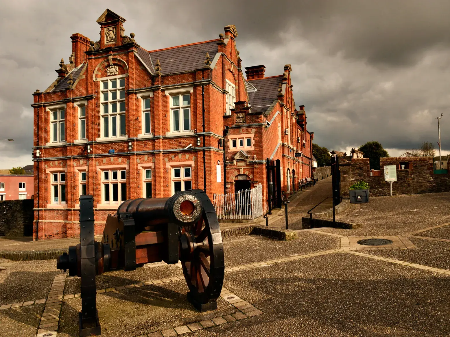 Historische Kanone auf dem Stadtwall von Londonderry mit Blick auf das rote Backsteingerichtsgebäude bei dramatischem Wolkenhimmel.
