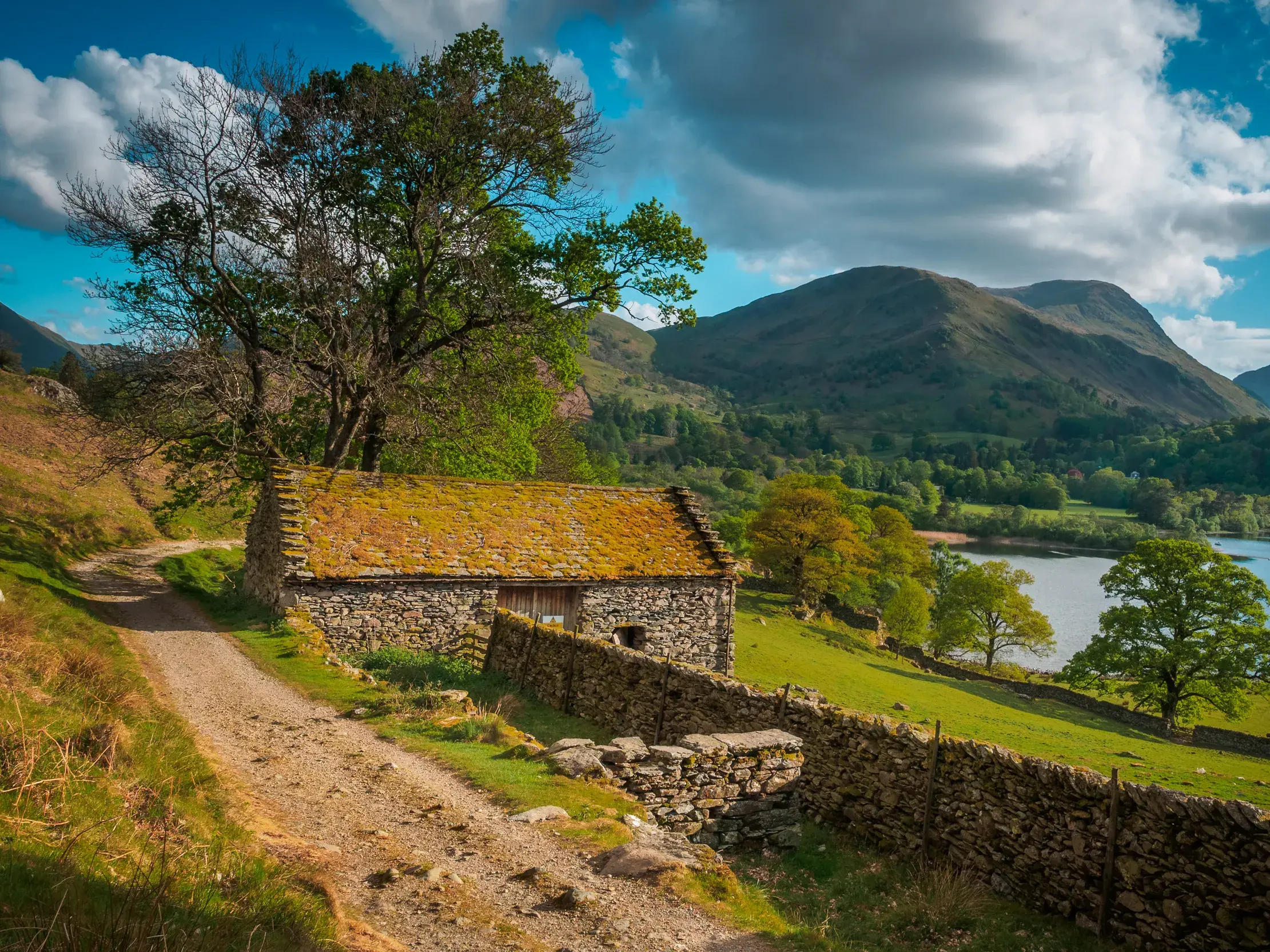 Ein Wanderpfad auf dem Coast to Coast Walk schlängelt sich an einer Steinhütte vorbei, mit Blick auf einen See im Lake District.