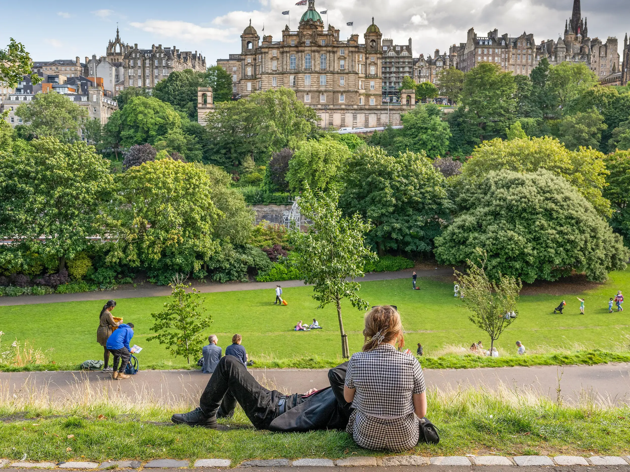 Menschen genießen einen sonnigen Tag in einem Park mit Blick auf die beeindruckende Architektur der Altstadt von Edinburgh.