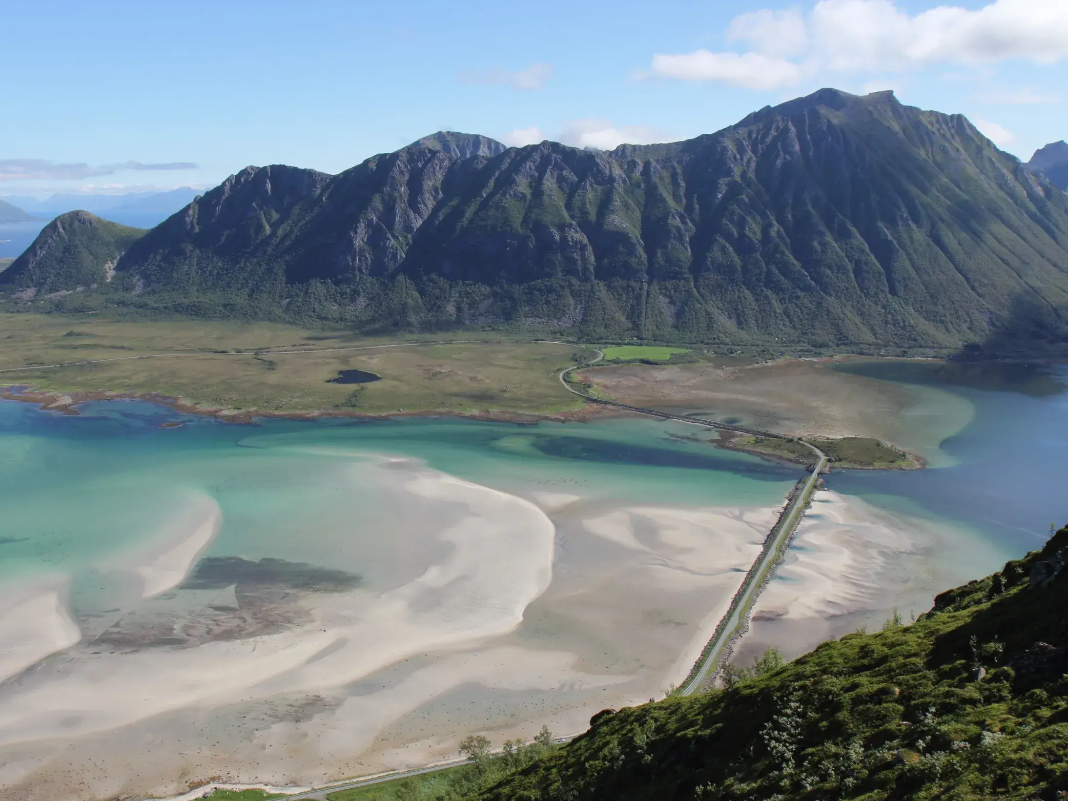 Wander-Aussicht vom Gipfel auf den Lofoten: türkiser Fjord mit Sandbänken, Brücke und eine schroffe Bergkette am Horizont.