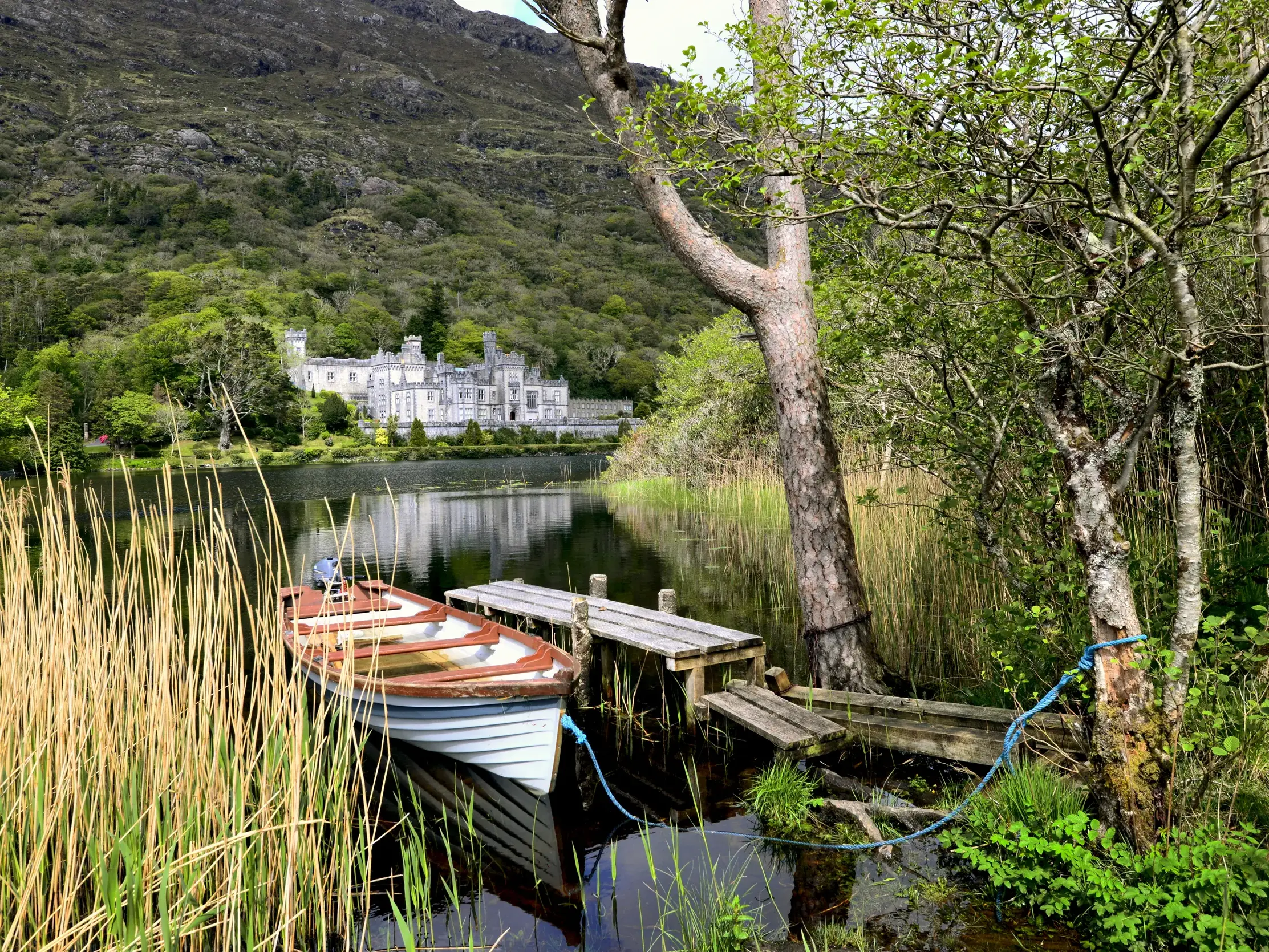 Ein Boot liegt an einem Holzsteg am Ufer, im Hintergrund die romantische Kylemore Abbey in der grünen Landschaft Connemaras.