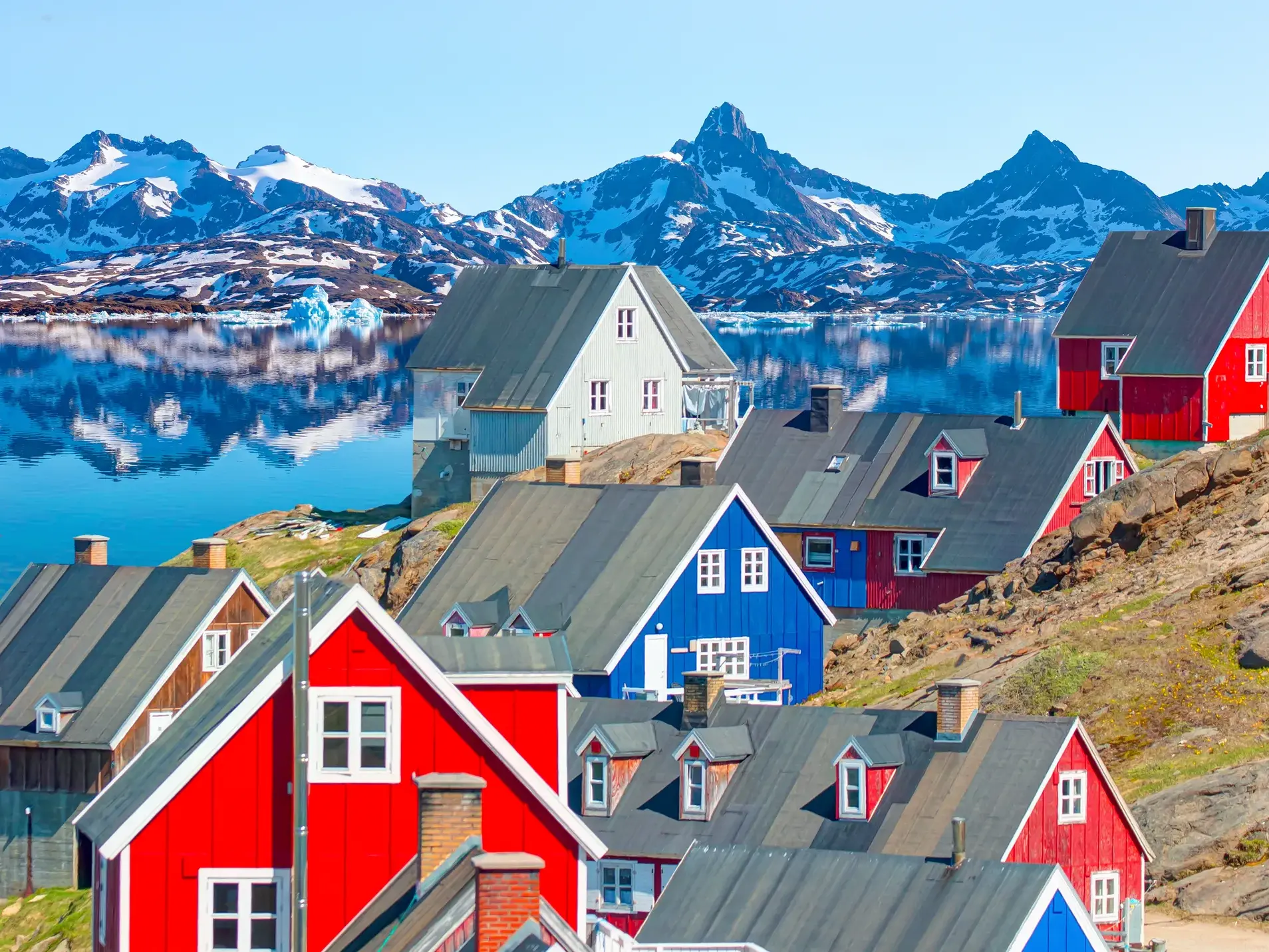 Bunte Holzhäuser am Hang in Tasiilaq mit Blick auf einen Fjord und die schneebedeckte Bergkulisse auf der Insel Ammassalik.