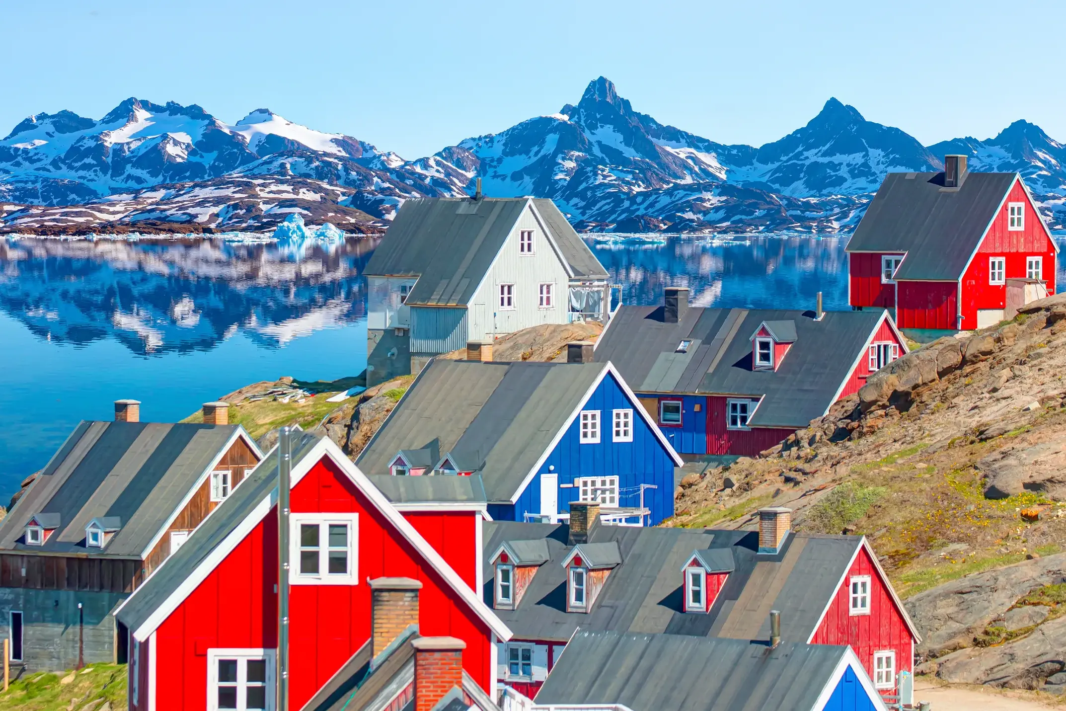 Bunte Holzhäuser am Hang in Tasiilaq mit Blick auf einen Fjord und die schneebedeckte Bergkulisse auf der Insel Ammassalik.