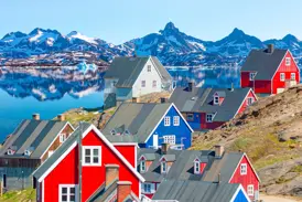 Bunte Holzhäuser am Hang in Tasiilaq mit Blick auf einen Fjord und die schneebedeckte Bergkulisse auf der Insel Ammassalik.